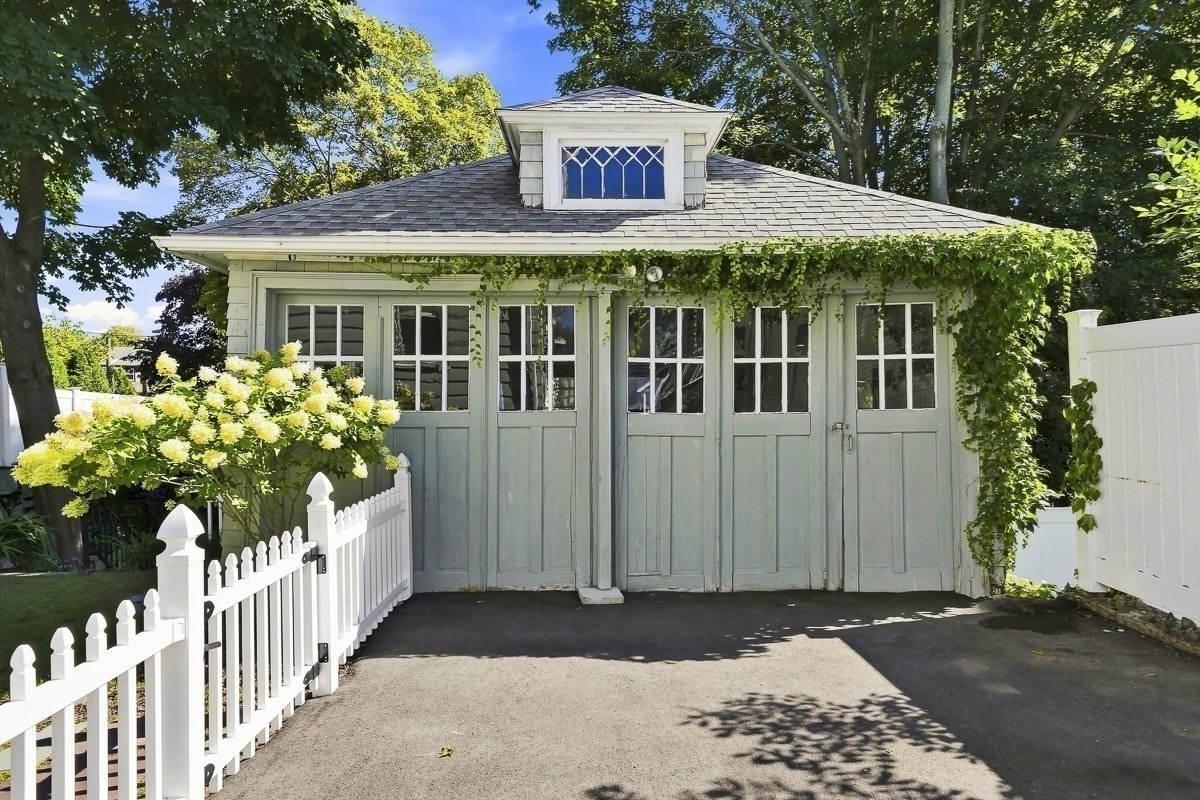 Sage-green detached garage with a decorative dormer, ivy-covered eaves, and white picket fence.