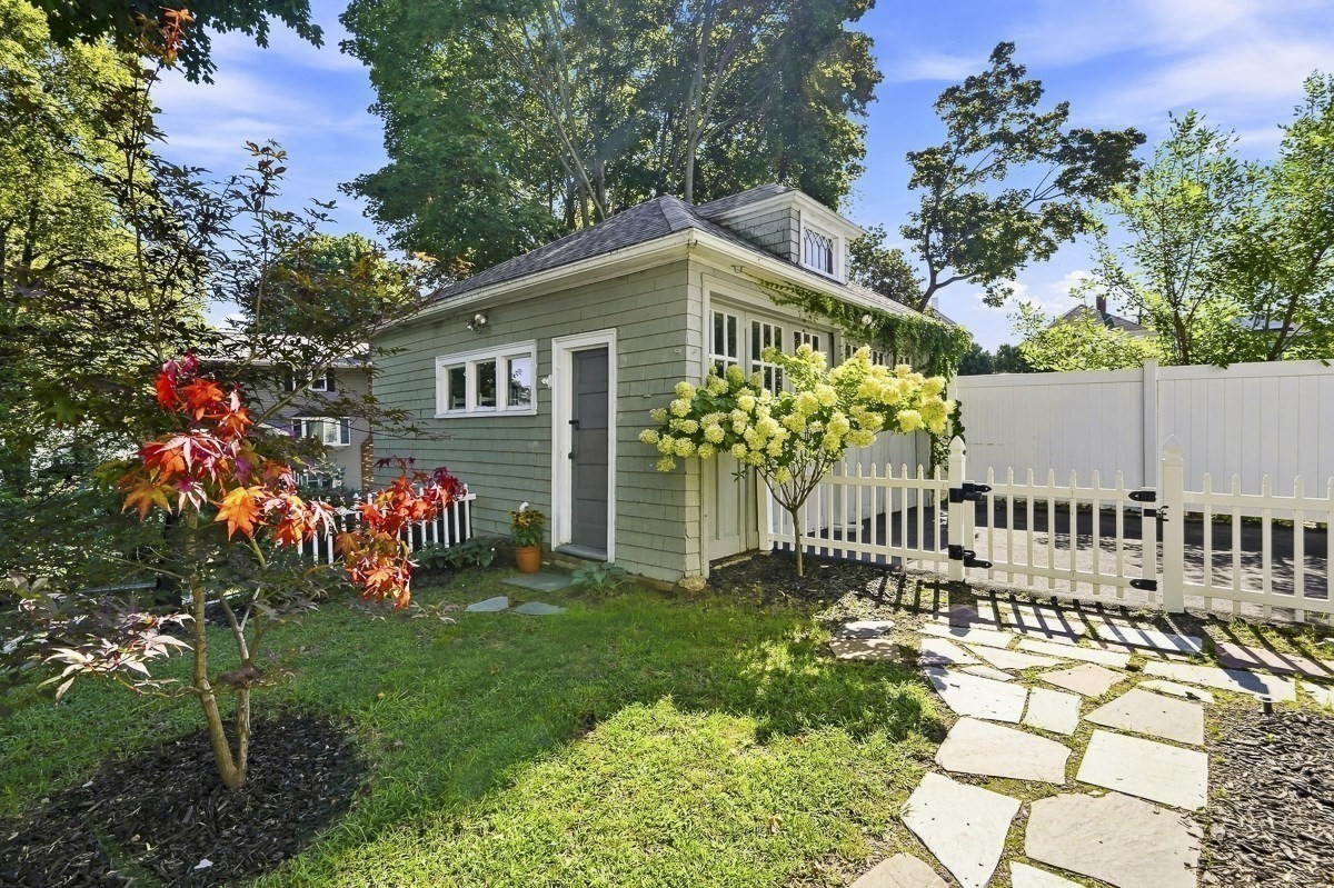 Green backyard outbuilding beside a white picket gate, flowering hydrangeas, and a stone path.