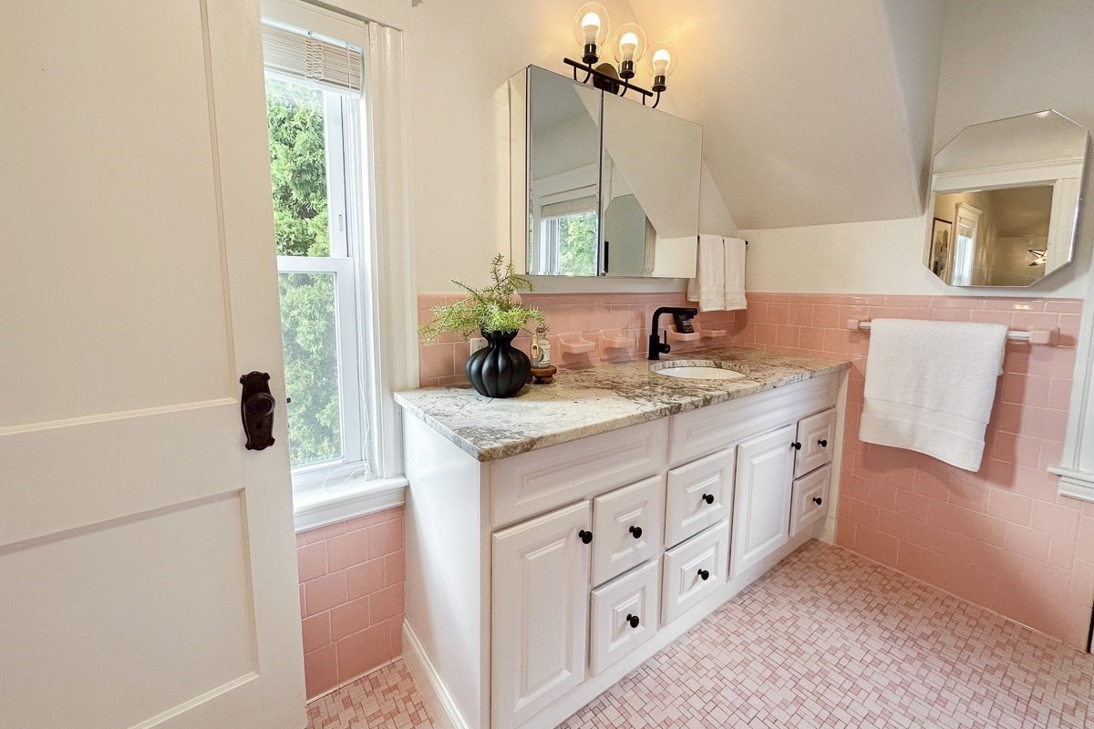 Bathroom vanity with white cabinetry, pink tile, mirrored cabinet, and a tall window.