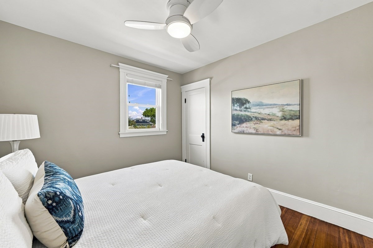 Staged bedroom with a white bed, blue accent pillow, ceiling fan, and hardwood floor.