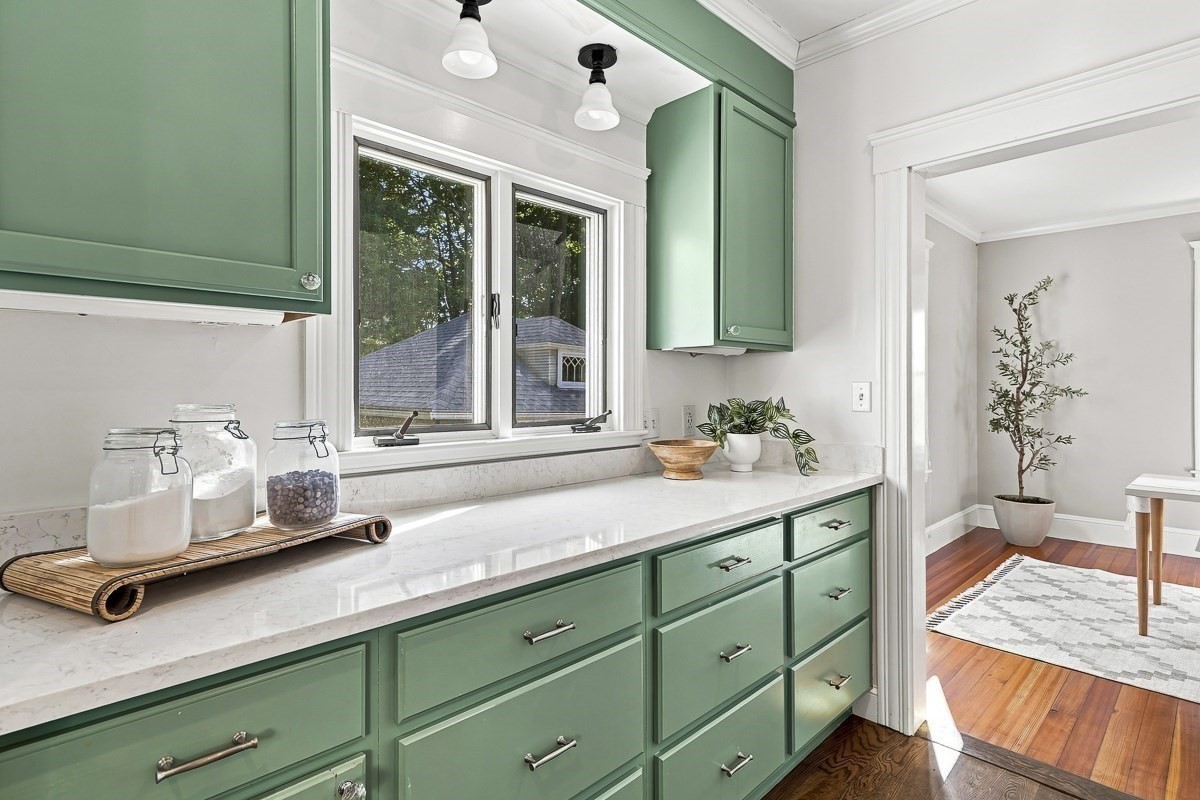 Kitchen with sage green cabinets, white stone counters, and a double window overlooking trees.