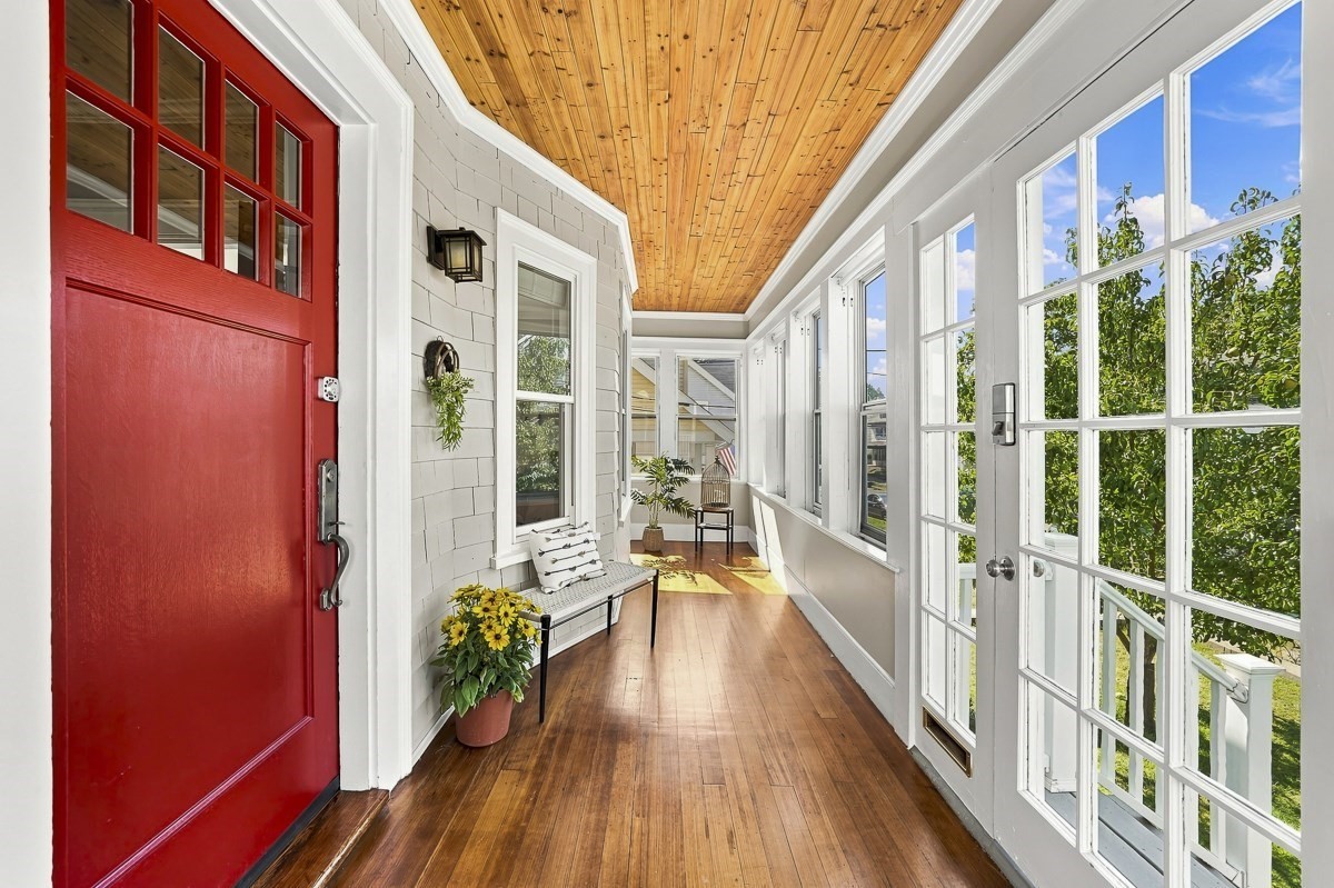 Enclosed sunroom with a red entry door, wood-plank ceiling, and broad windows overlooking greenery.