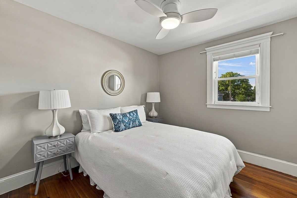 Staged bedroom with a white bed, ceiling fan, and window overlooking leafy trees.