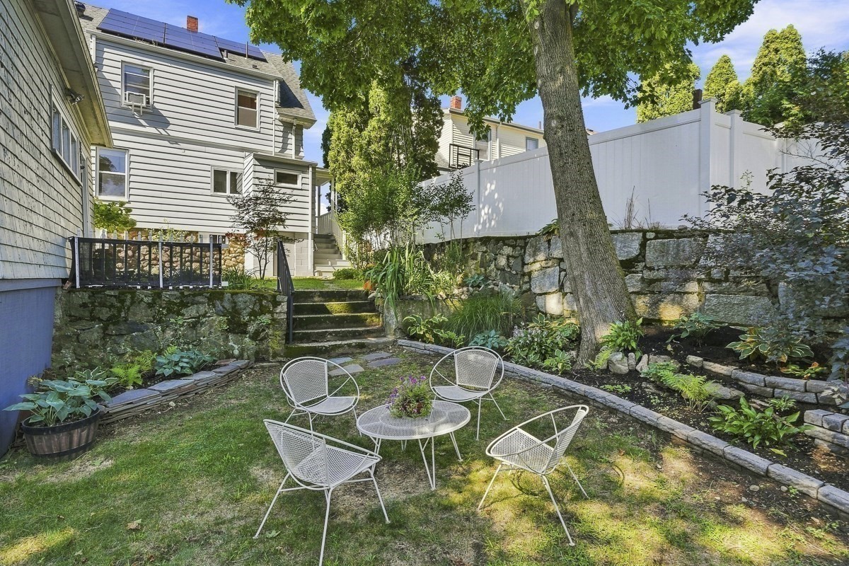 Backyard patio with white metal seating, stone garden walls, steps, and a tall privacy fence.