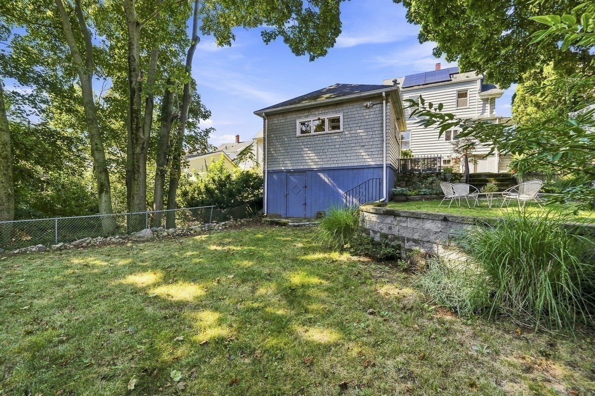 Backyard with a gray outbuilding, mature trees, and a raised patio with outdoor chairs.