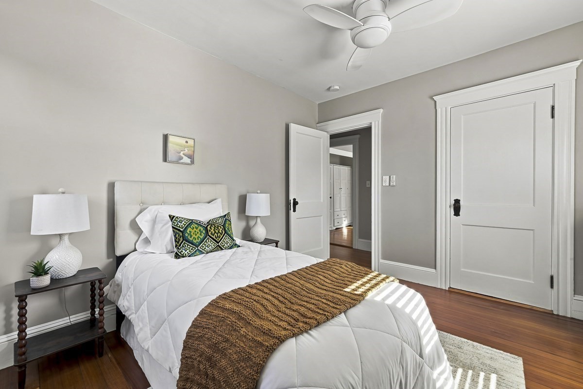 Staged bedroom with a white bed, patterned pillows, brown knit throw, and hardwood floors.
