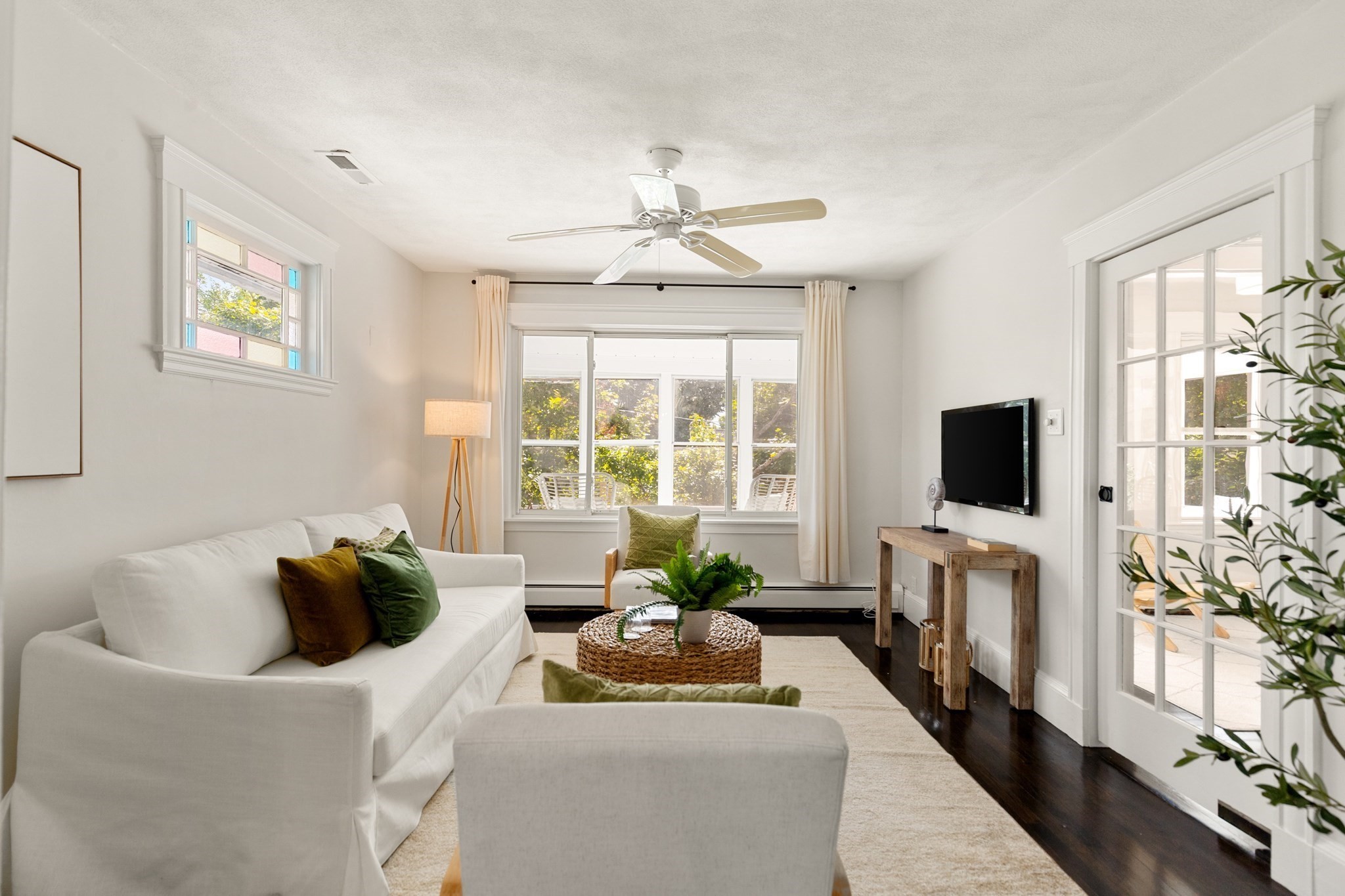 Staged living room with white sofas, a woven coffee table, and windows overlooking greenery.