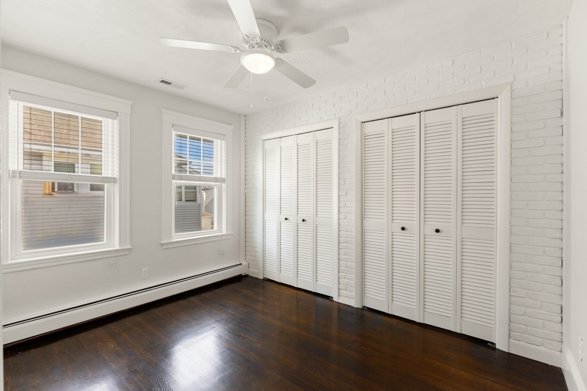 Empty bedroom with dark hardwood floors, white brick wall, louvered closets, and two windows.