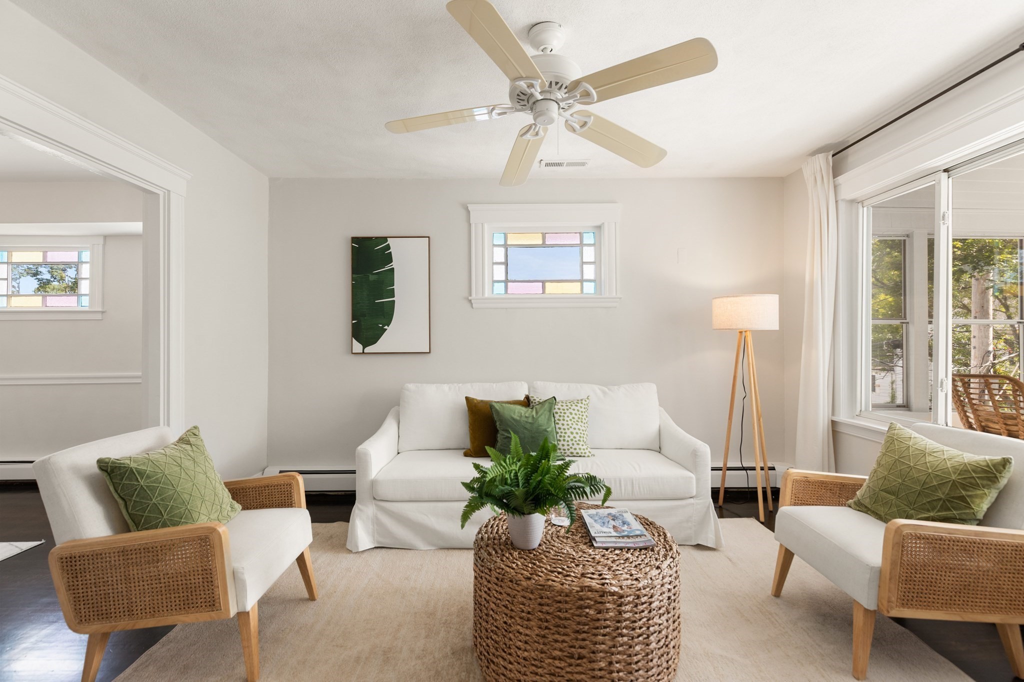 Staged living room with white seating, green accents, a woven coffee table, and large windows.