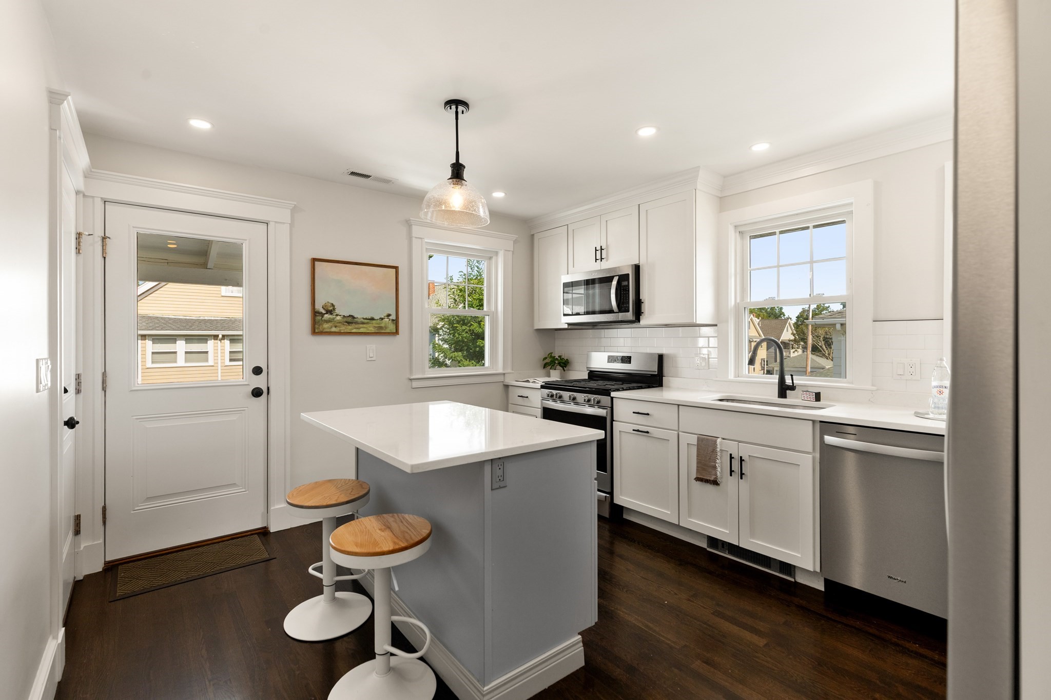 Bright white kitchen with a gray island, wooden stools, stainless steel appliances, and dark hardwood floors.
