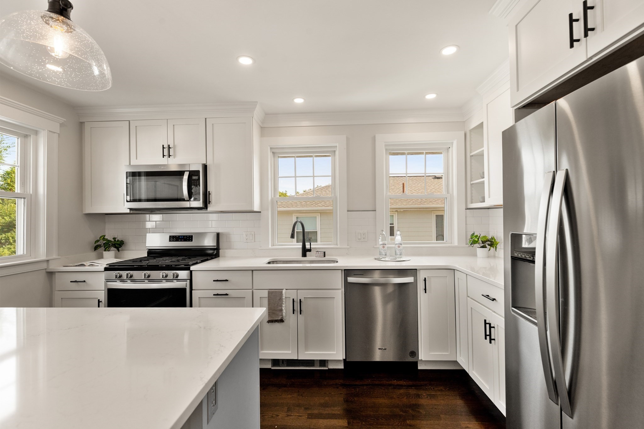 Bright white kitchen with a large stone island, stainless steel appliances, and windows above the sink.