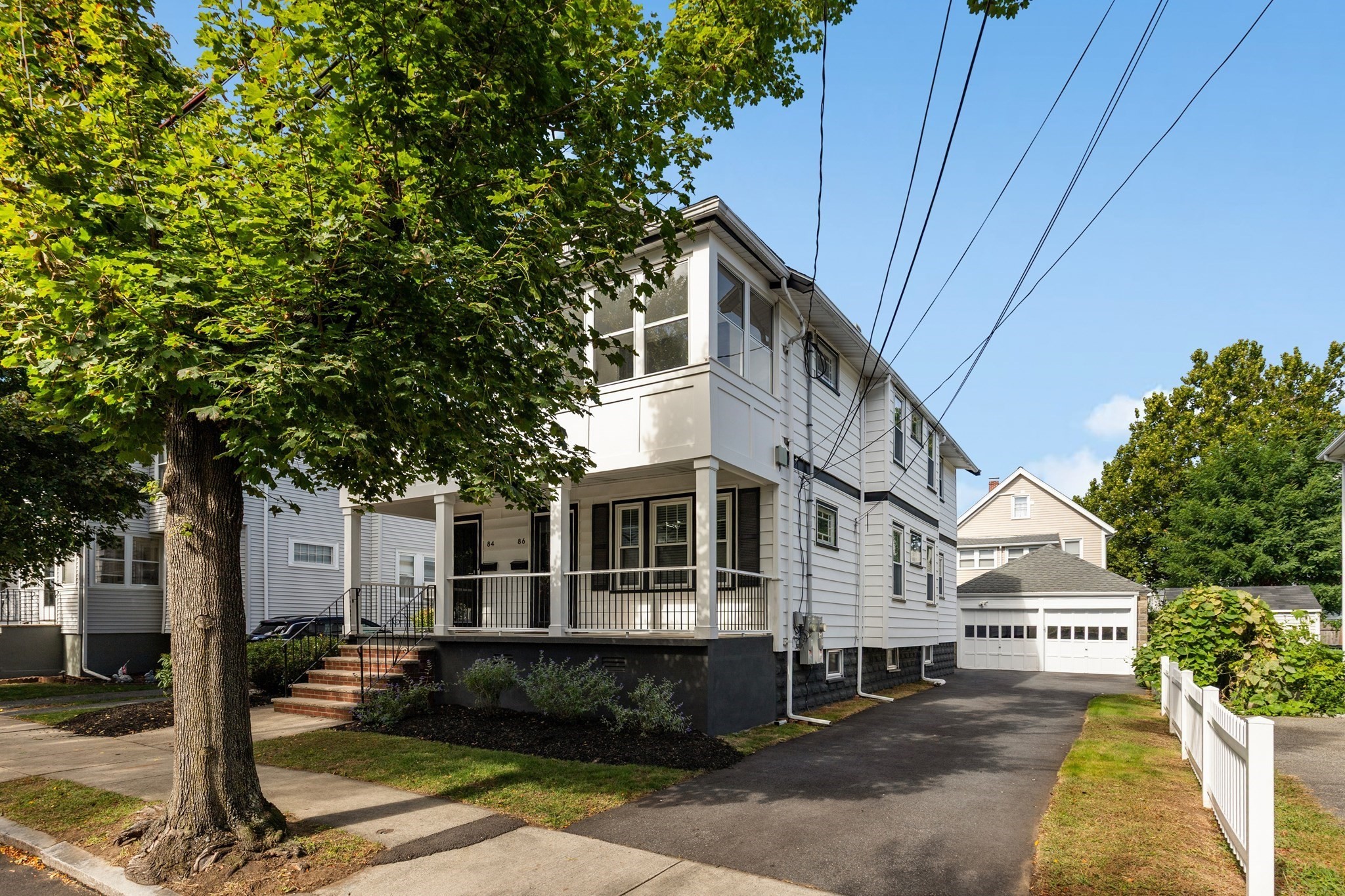 White multi-unit home with a raised porch, driveway, detached garage, and large leafy tree.