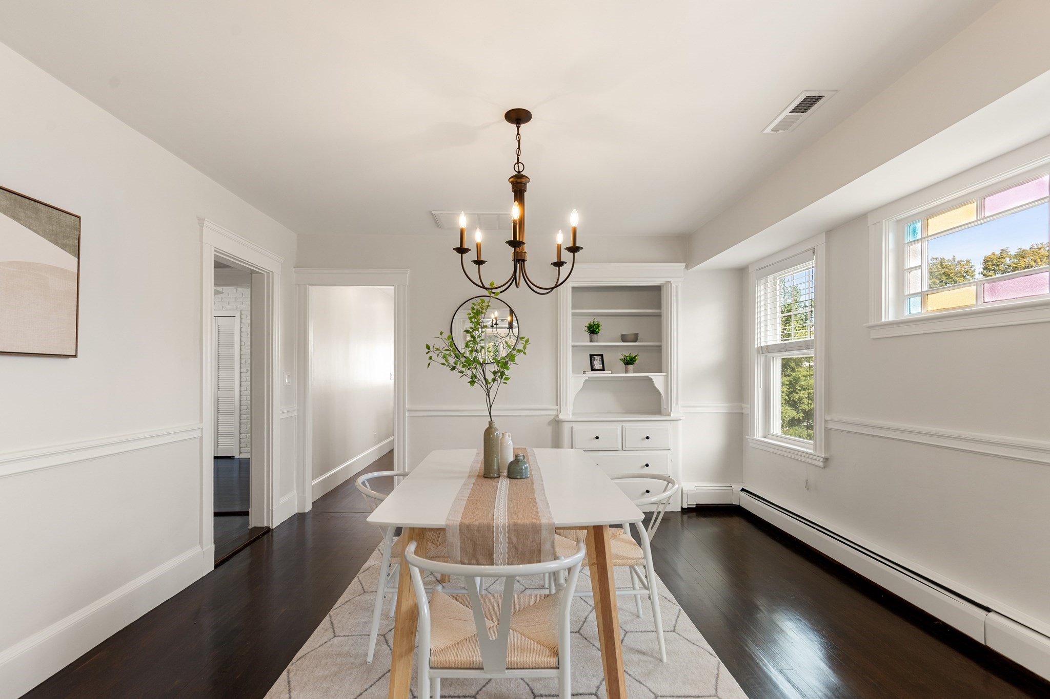 Bright dining room with a white table, dark hardwood floors, built-in shelves, and colorful window glass.
