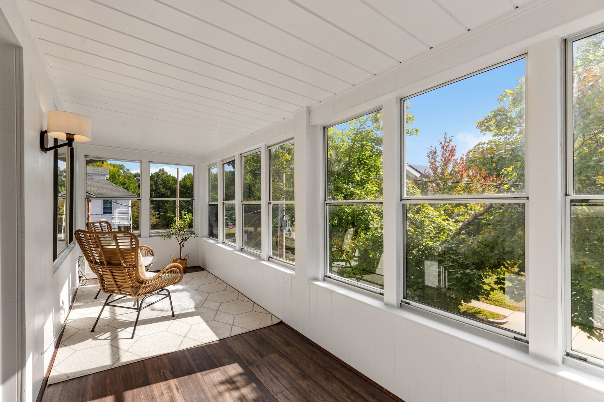 Furnished sunroom with wicker chairs, dark wood flooring, and large windows overlooking trees.
