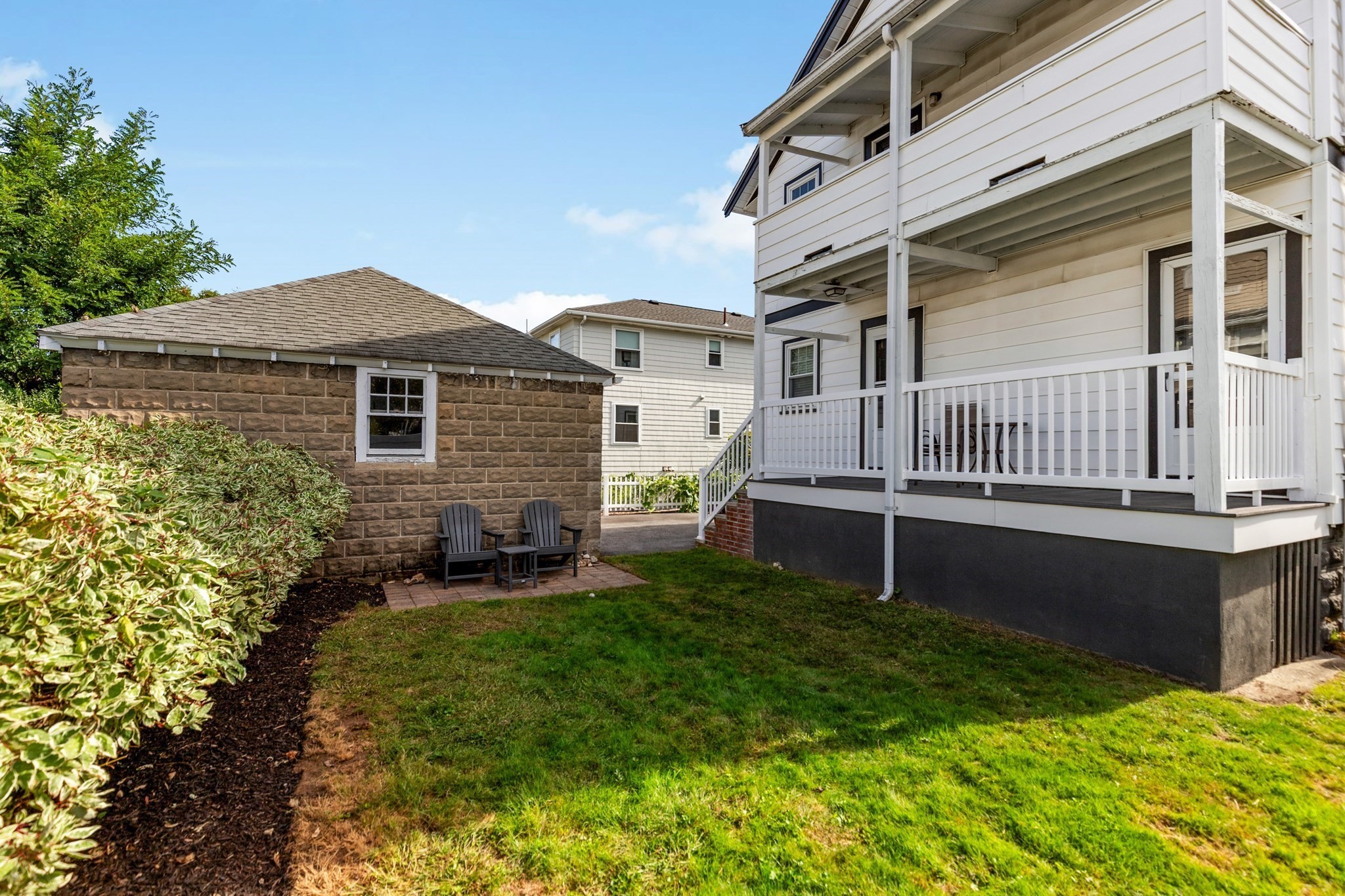 Home’s backyard with a covered porch, green lawn, and stone outbuilding with patio chairs.