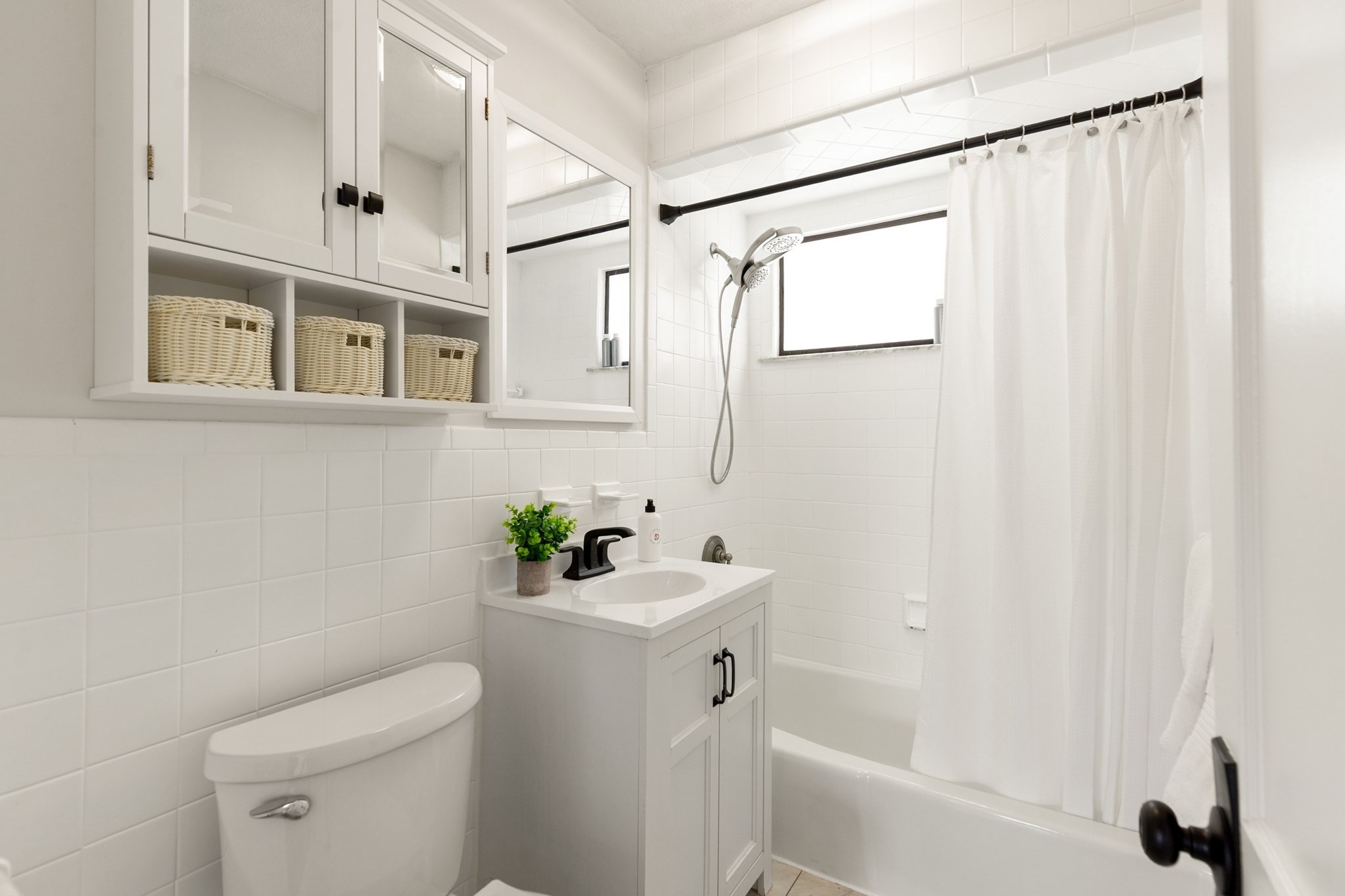 White-tiled bathroom with mirrored storage, a vanity, toilet, and tub-shower behind a curtain.