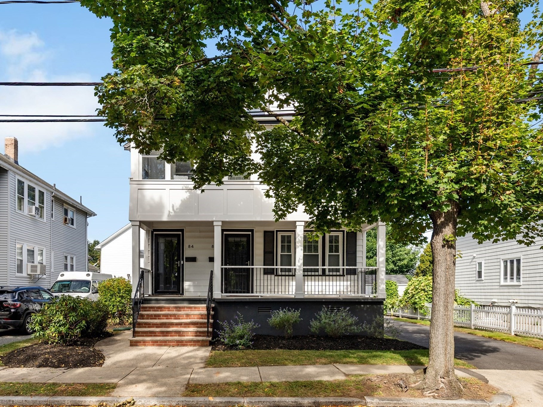 White two-story residence with a covered porch and brick front steps beneath a large leafy tree.