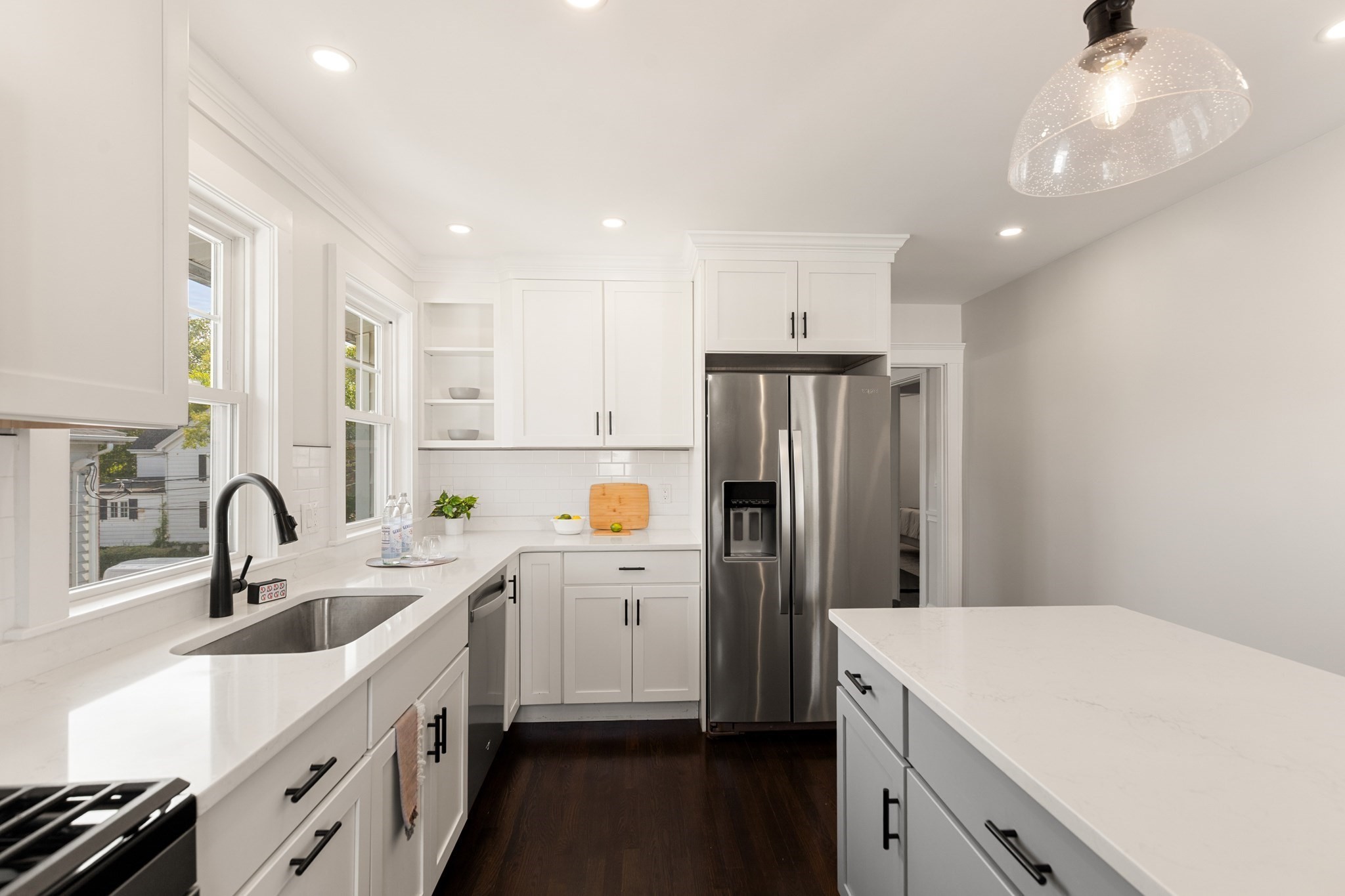 Bright white kitchen with a large island, stainless steel refrigerator, and sink beneath two windows.
