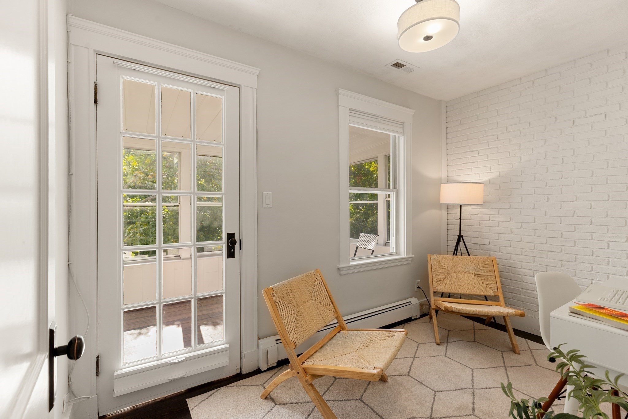Sitting area with woven chairs, a white brick wall, and a glass-paneled door to a leafy outdoor space.