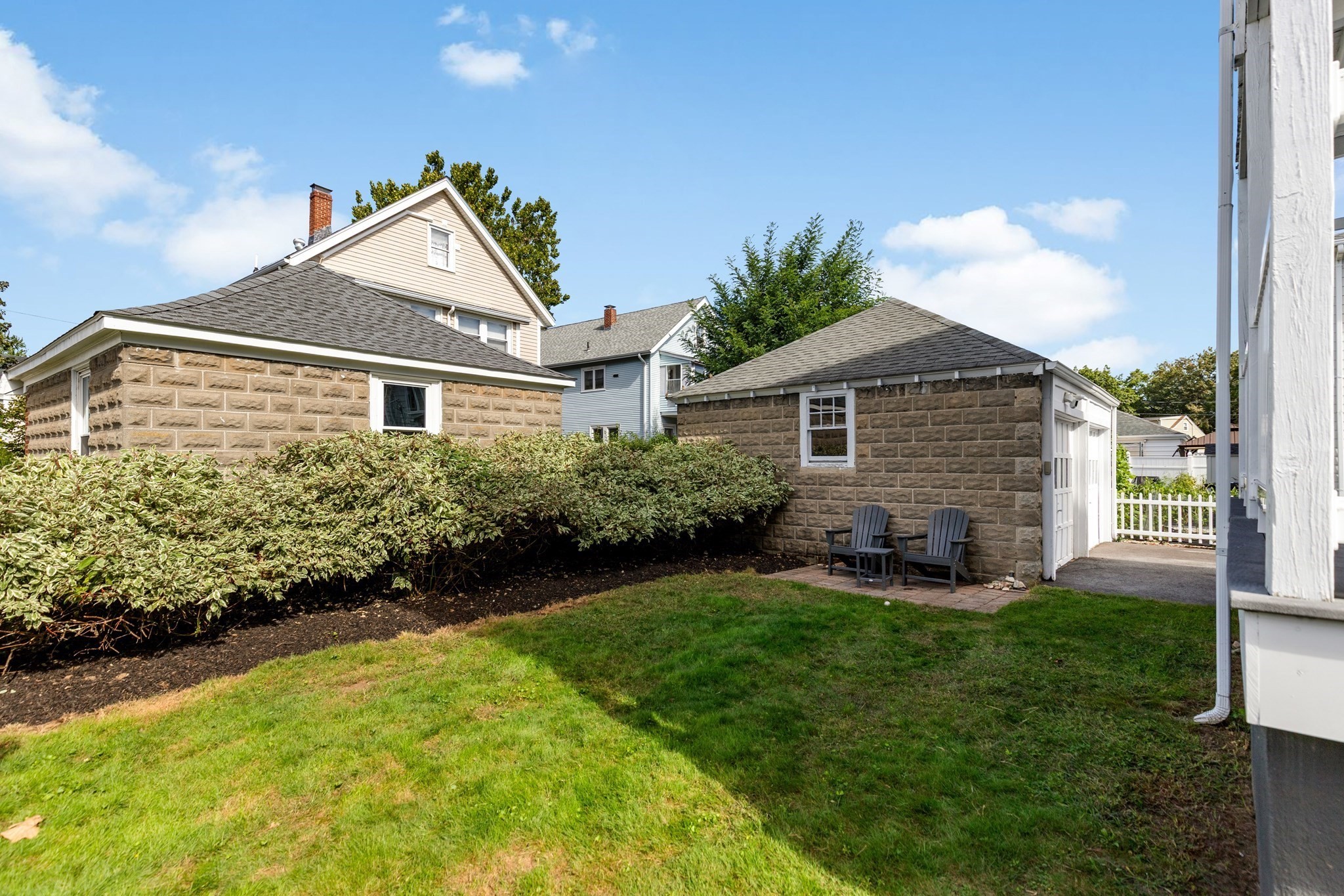 Backyard with lawn, hedges, stone outbuildings, and two chairs beside one building.