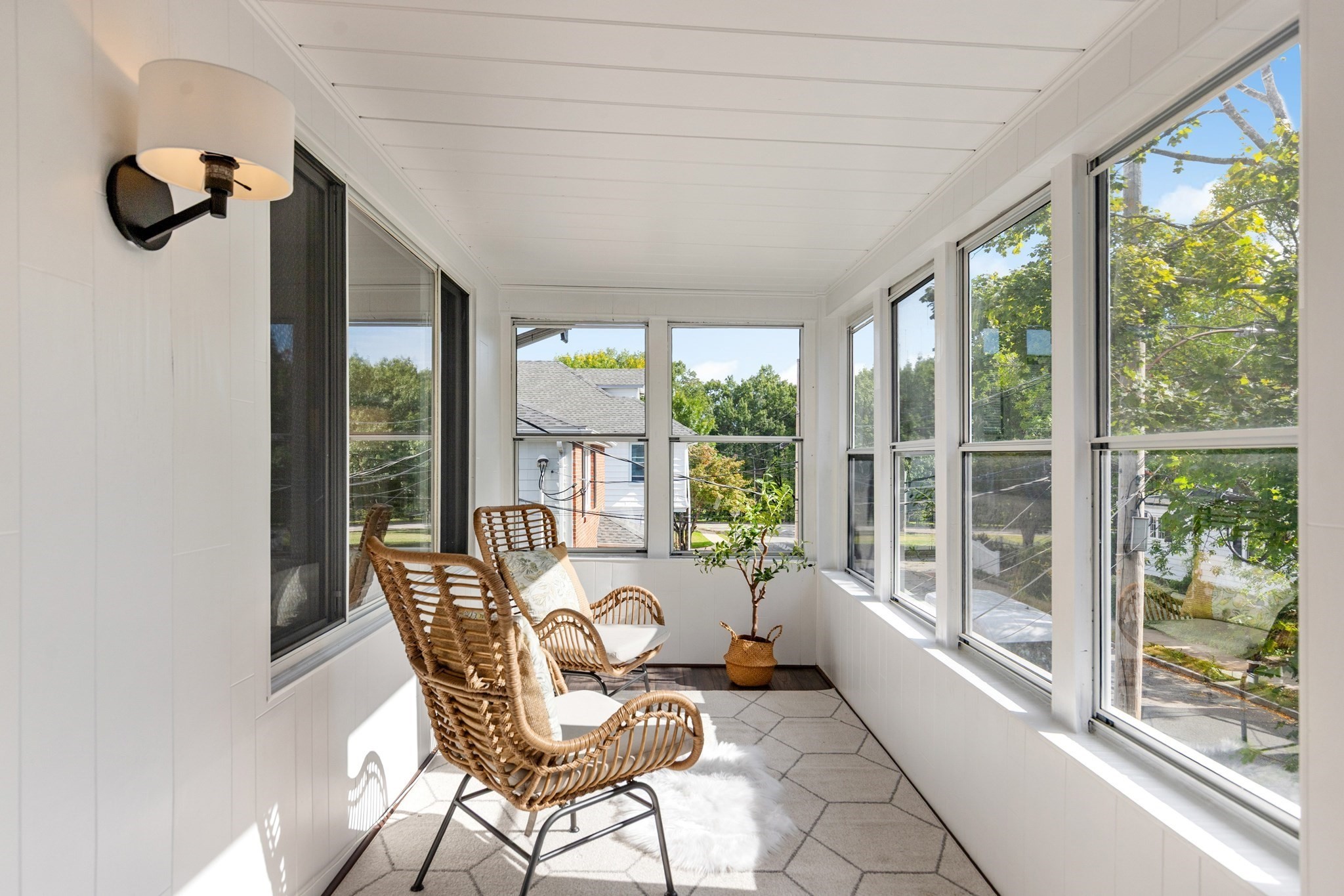 Bright sunroom with wicker chairs, a potted plant, and large windows overlooking leafy trees.