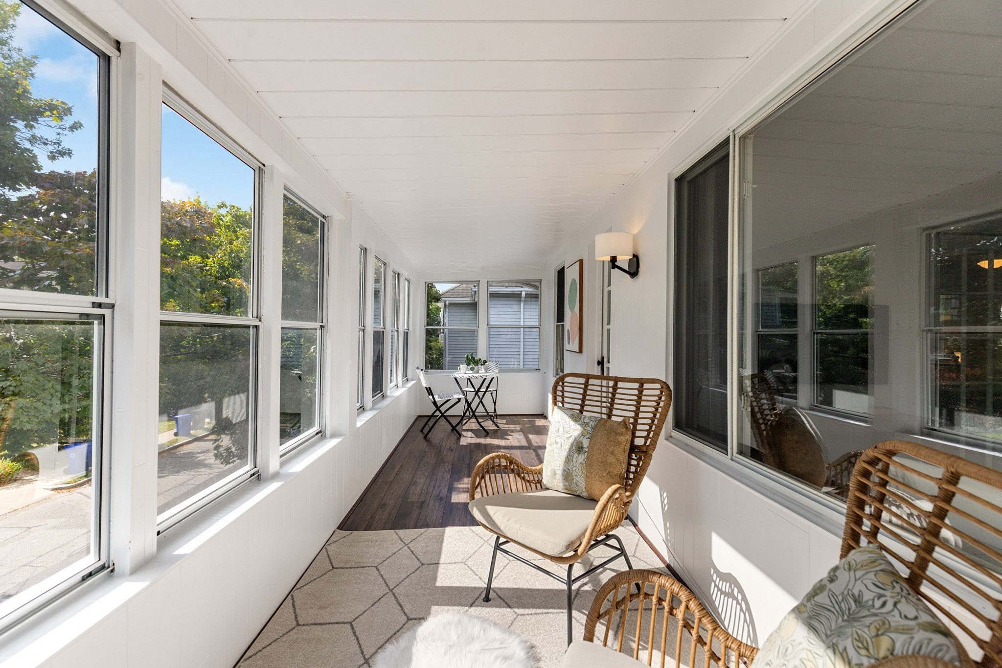 Bright enclosed sunroom with large windows, wicker seating, a café table, and dark wood flooring.