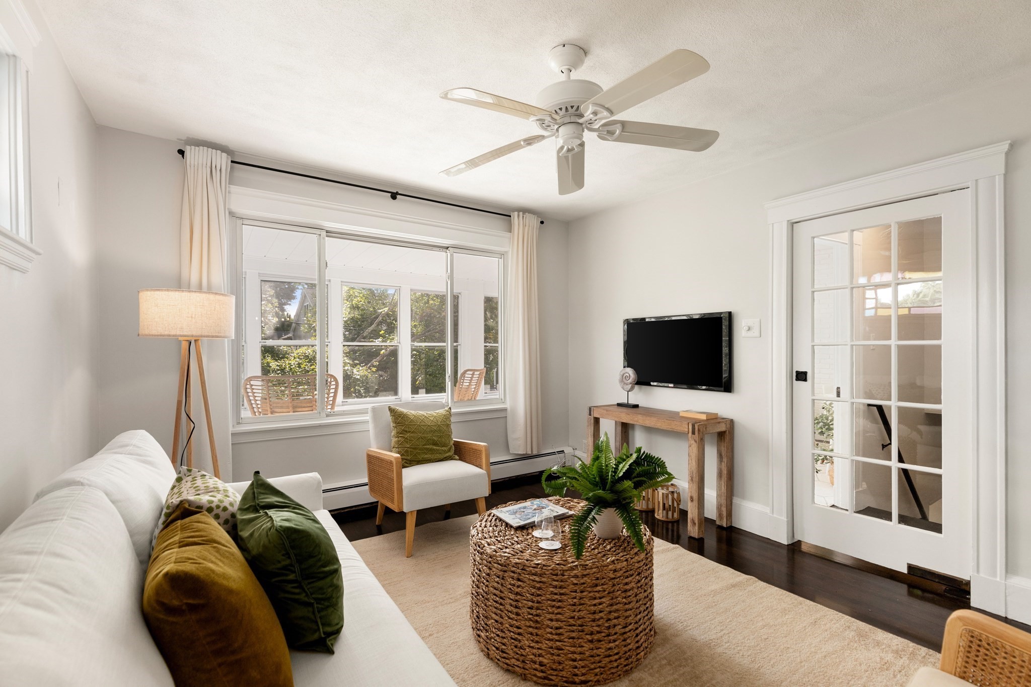 Bright living room with a white sofa, woven coffee table, and large windows overlooking trees.
