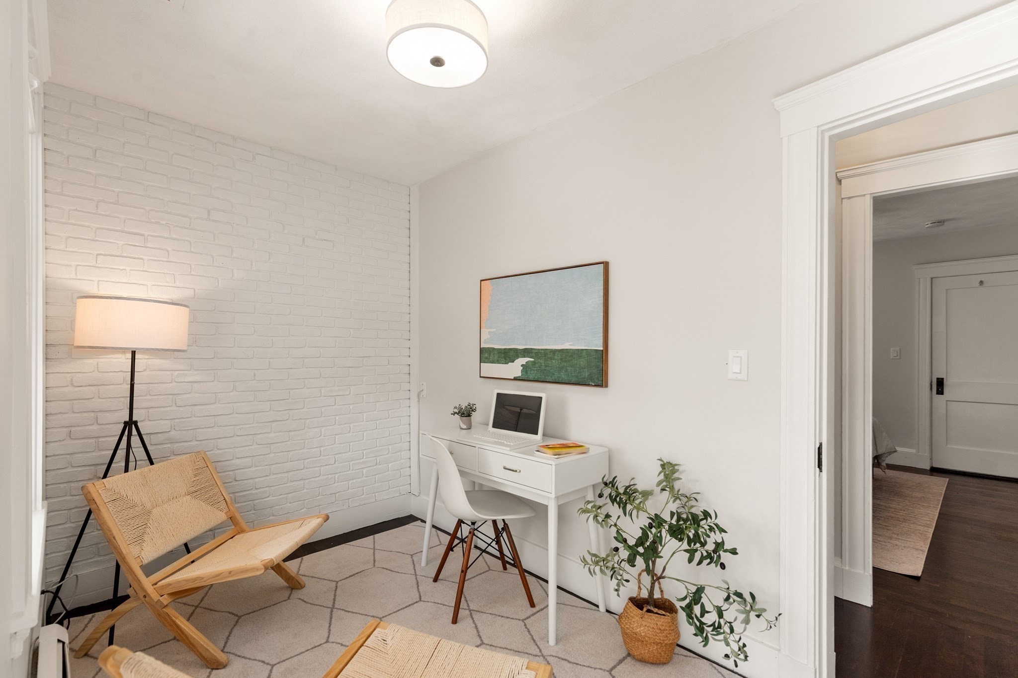 Staged home office with a white desk, brick accent wall, woven chair, and potted plants.