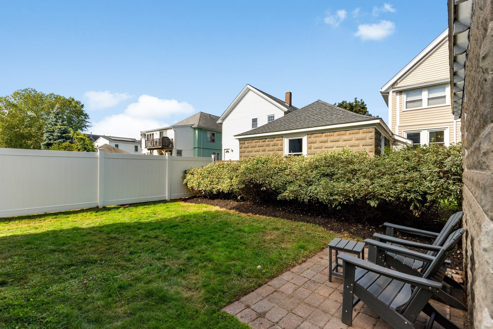 Backyard with a fenced lawn, hedge, and paved patio seating beside the house.