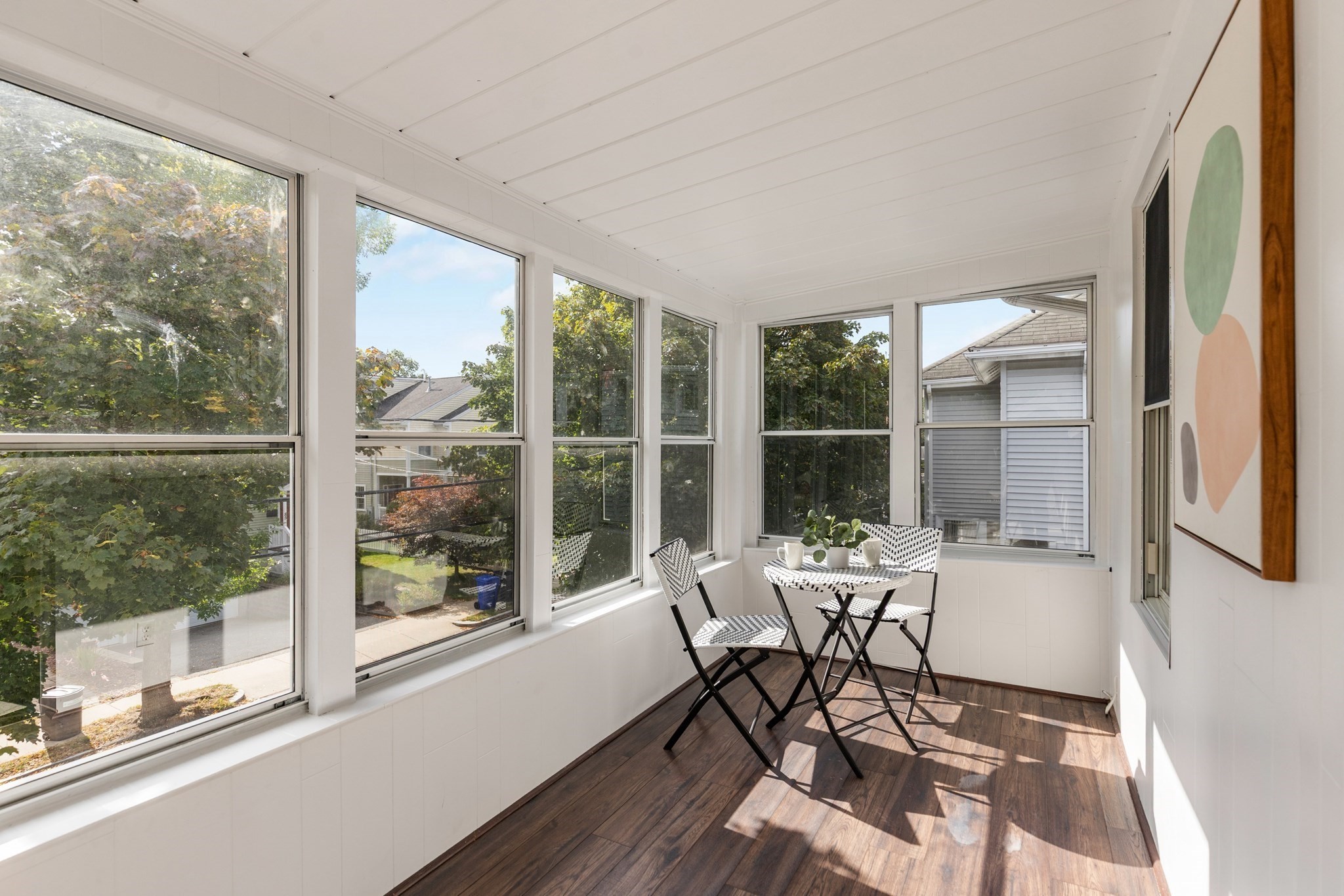 Bright enclosed sunroom with wraparound windows, dark wood floors, and a small black-and-white dining set.