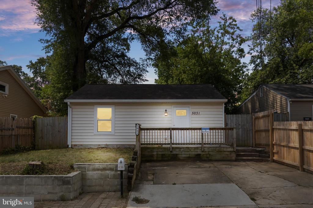 Single-story house with a raised wooden porch, concrete driveway, and fenced yard beneath mature trees.