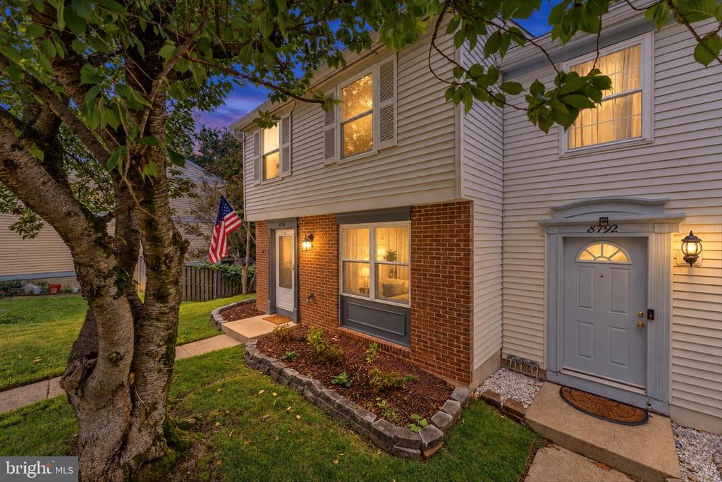 Two-story home with white siding, brick facade, illuminated windows, and a large tree in front.