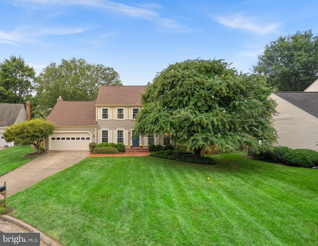 Two-story beige house with an attached garage, wide lawn, and mature trees.