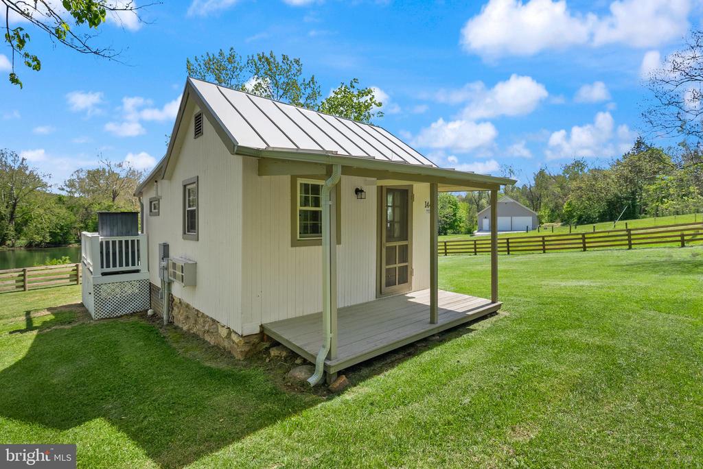 Small white cottage with a metal roof and covered porch beside a pond.