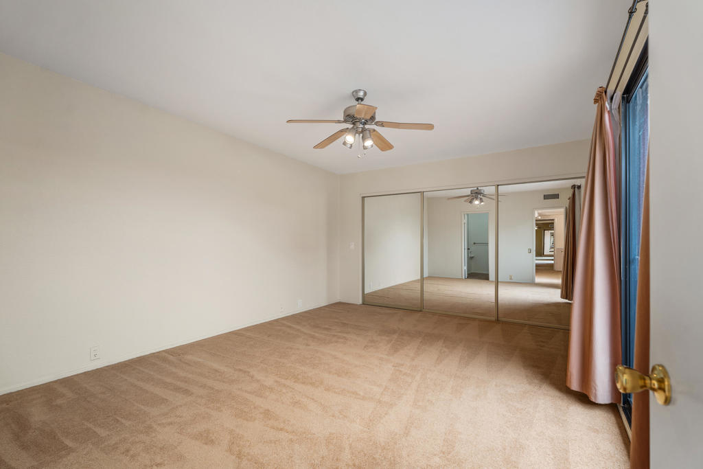 Empty bedroom with mirrored closet doors, beige carpet, and a ceiling fan.