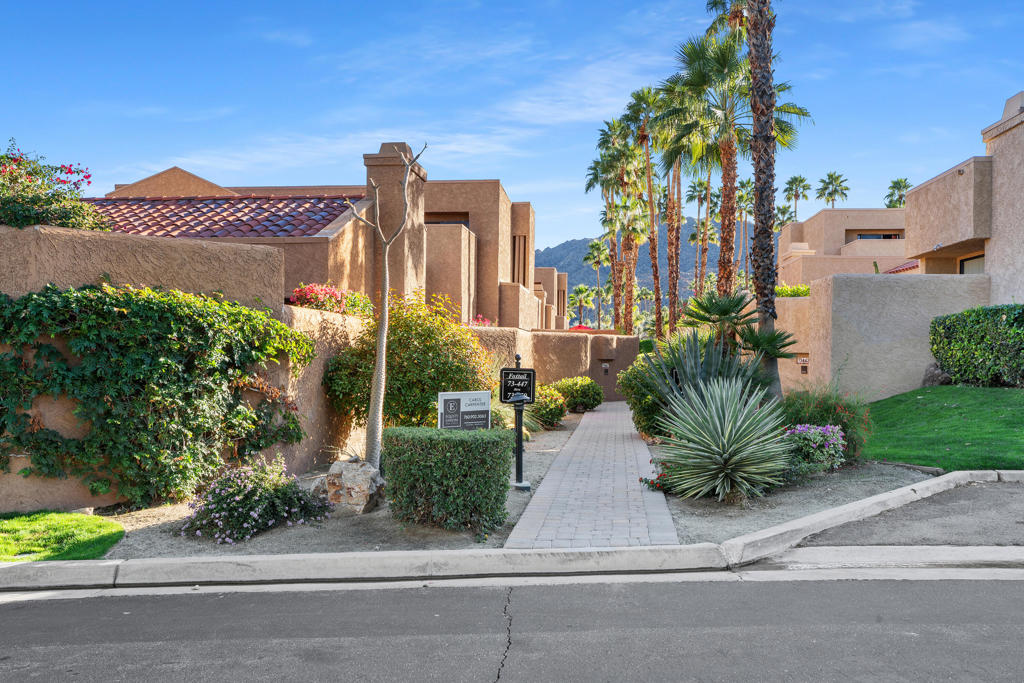 Residential community entrance with a paved walkway, desert landscaping, tall palms, and mountains beyond.