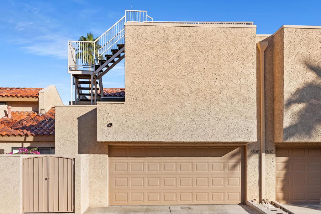 Beige stucco home with a paneled garage, front gate, and exterior stairs to a rooftop terrace.