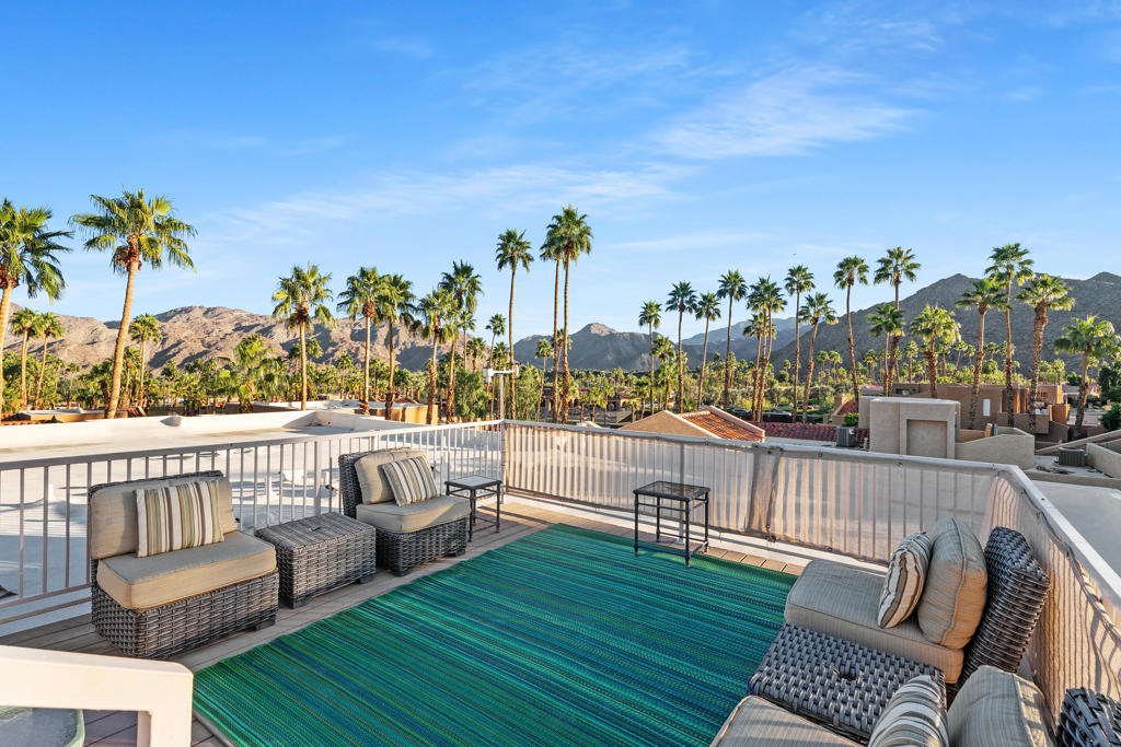Rooftop terrace with wicker seating and a turquoise rug overlooking palm trees and mountains.
