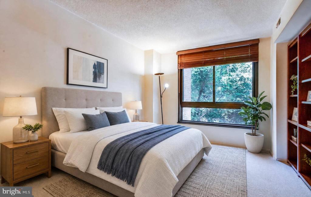 Staged bedroom with a white bed, charcoal throw, and a wide window overlooking leafy trees.