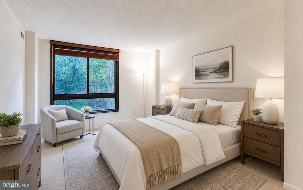 Staged bedroom with a king-size bed, wood nightstands, and a large window overlooking trees.