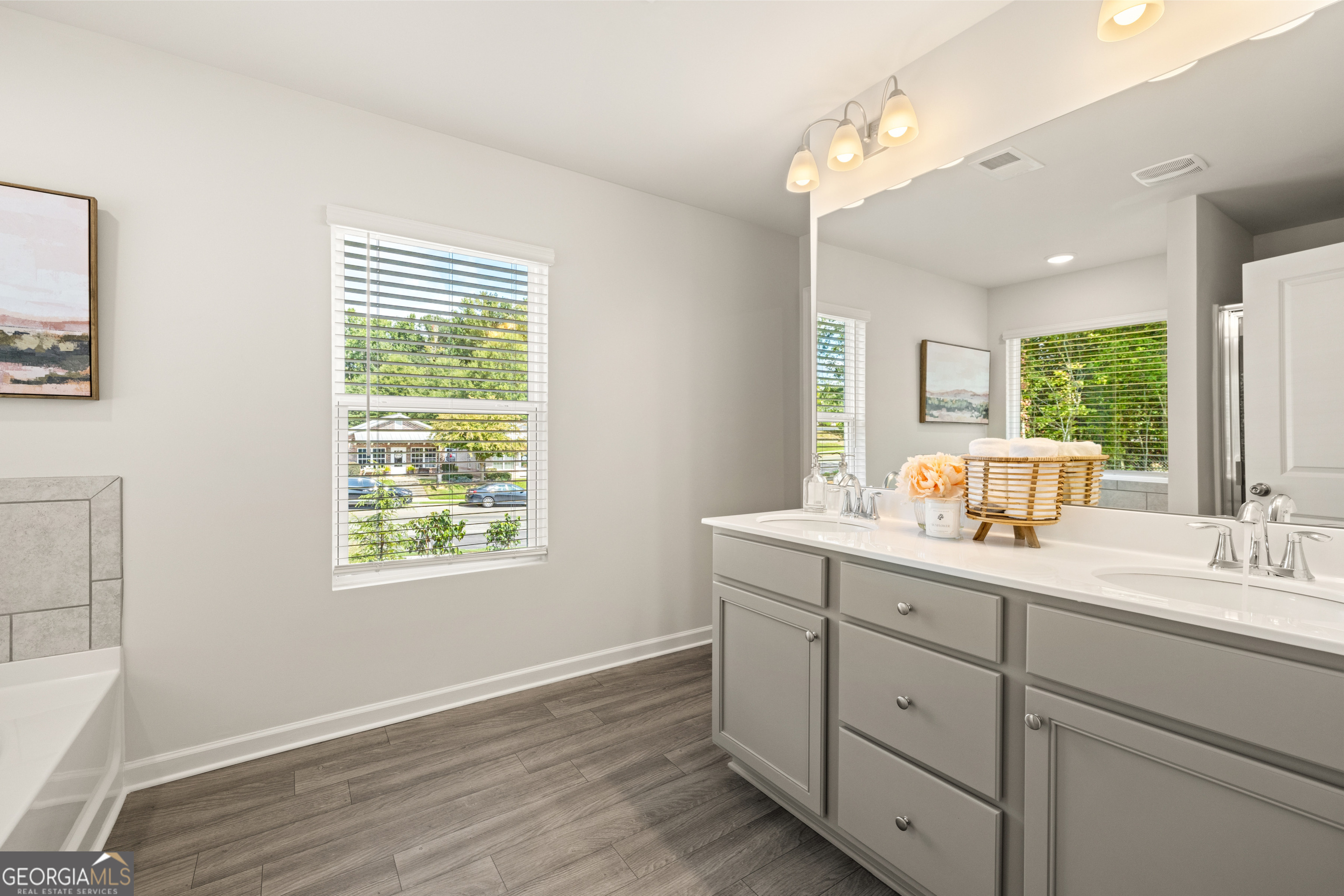 Staged bathroom with a gray double-sink vanity, large mirror, and wood-look flooring.