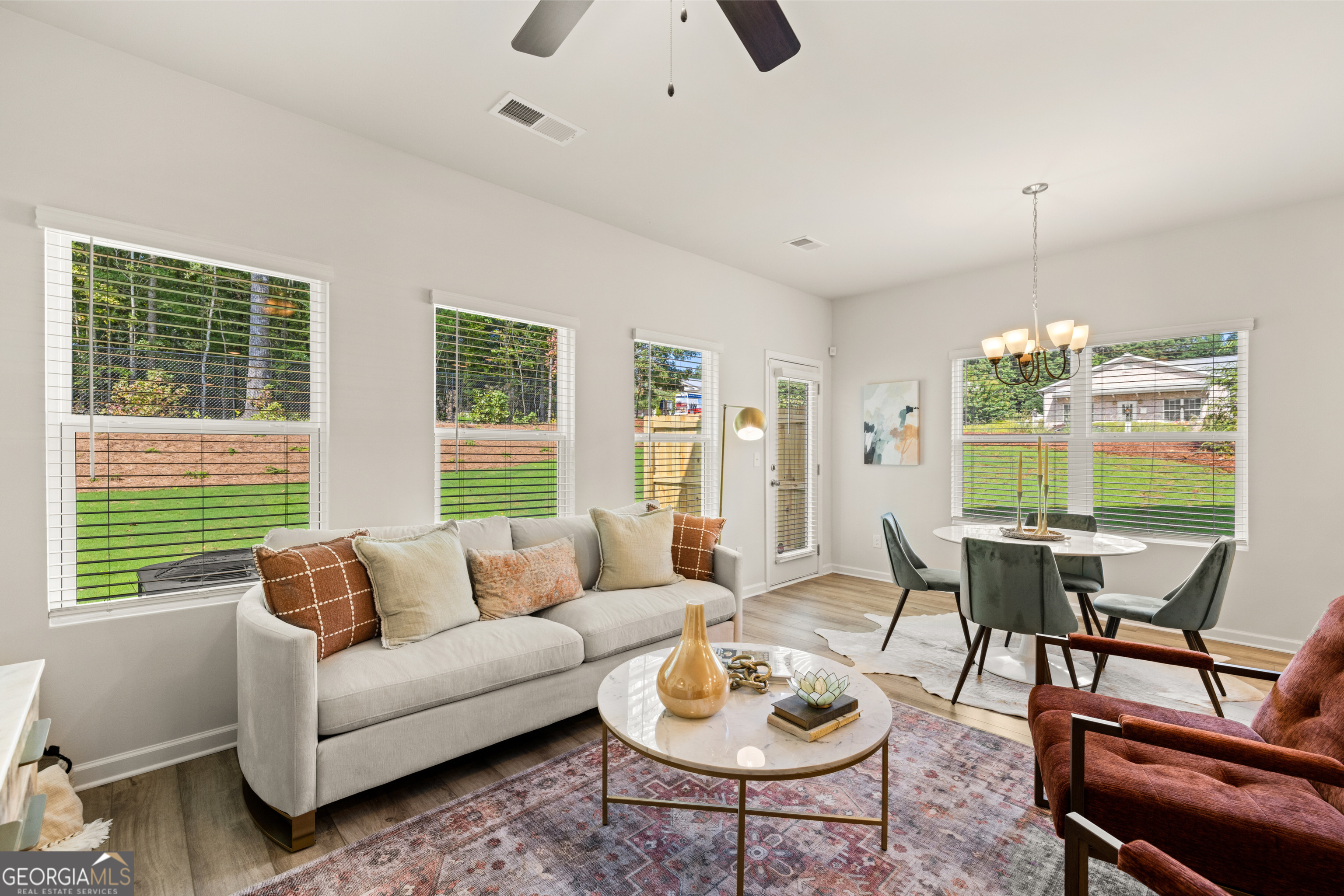 Staged living and dining room with a gray sofa, rust armchairs, and large windows overlooking a green lawn.