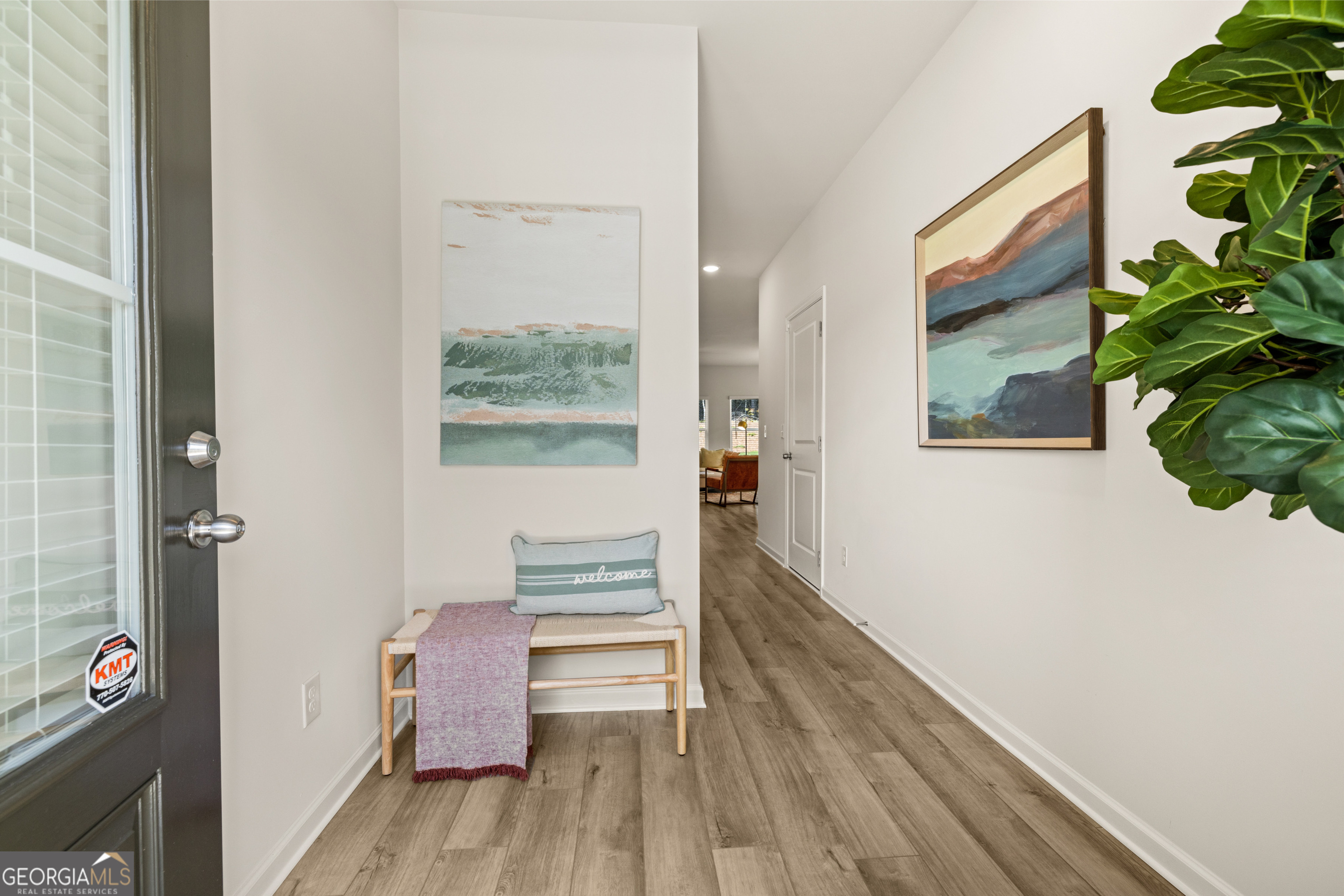 Staged entryway with a bench, welcome pillow, wood-look floors, wall art, and a leafy plant.