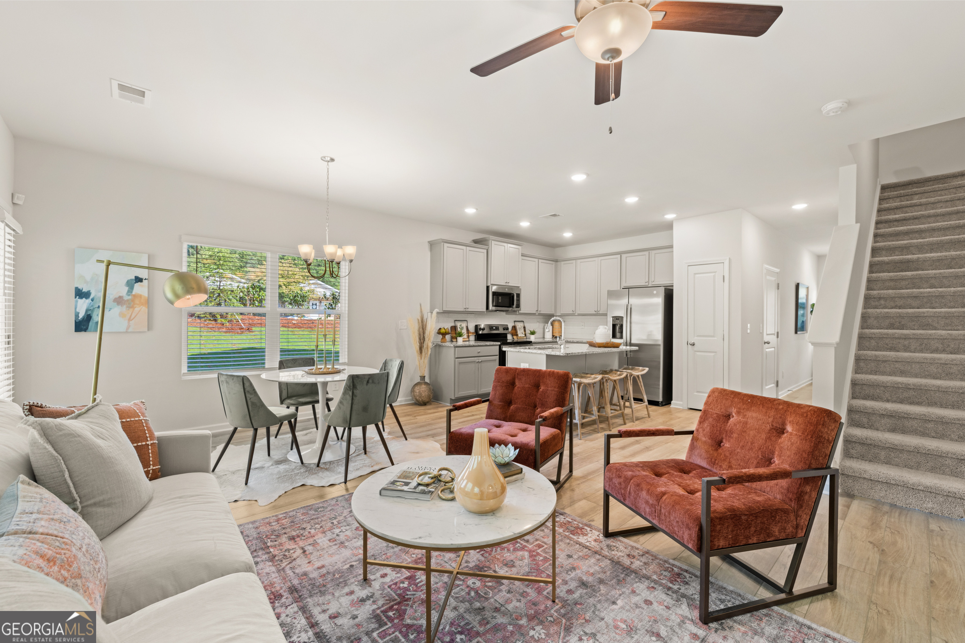 Open-plan living room, dining area, and kitchen with gray cabinets, rust-colored armchairs, and carpeted stairs.