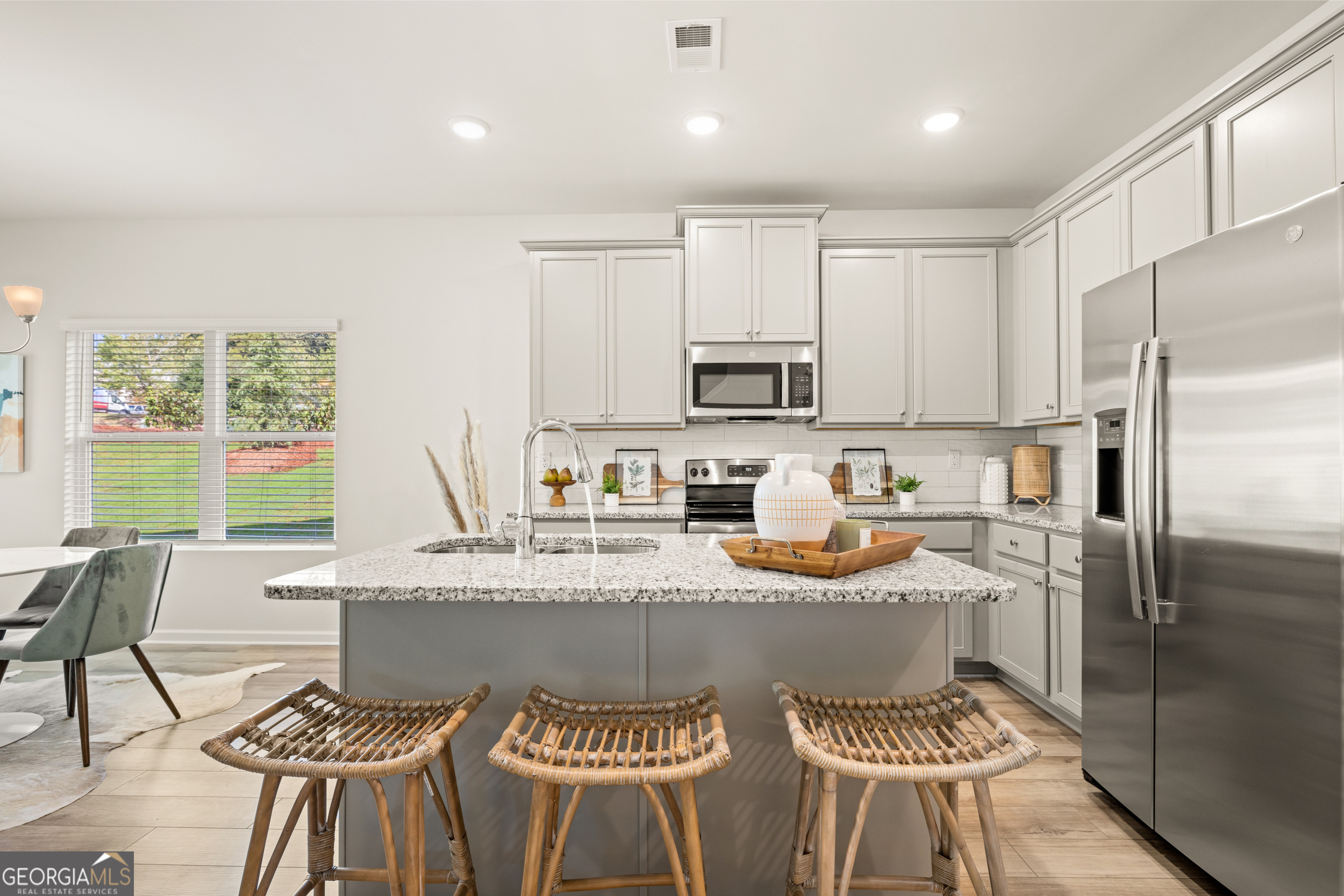 Kitchen with a large gray island, speckled stone countertop, white cabinets, and three woven bar stools.