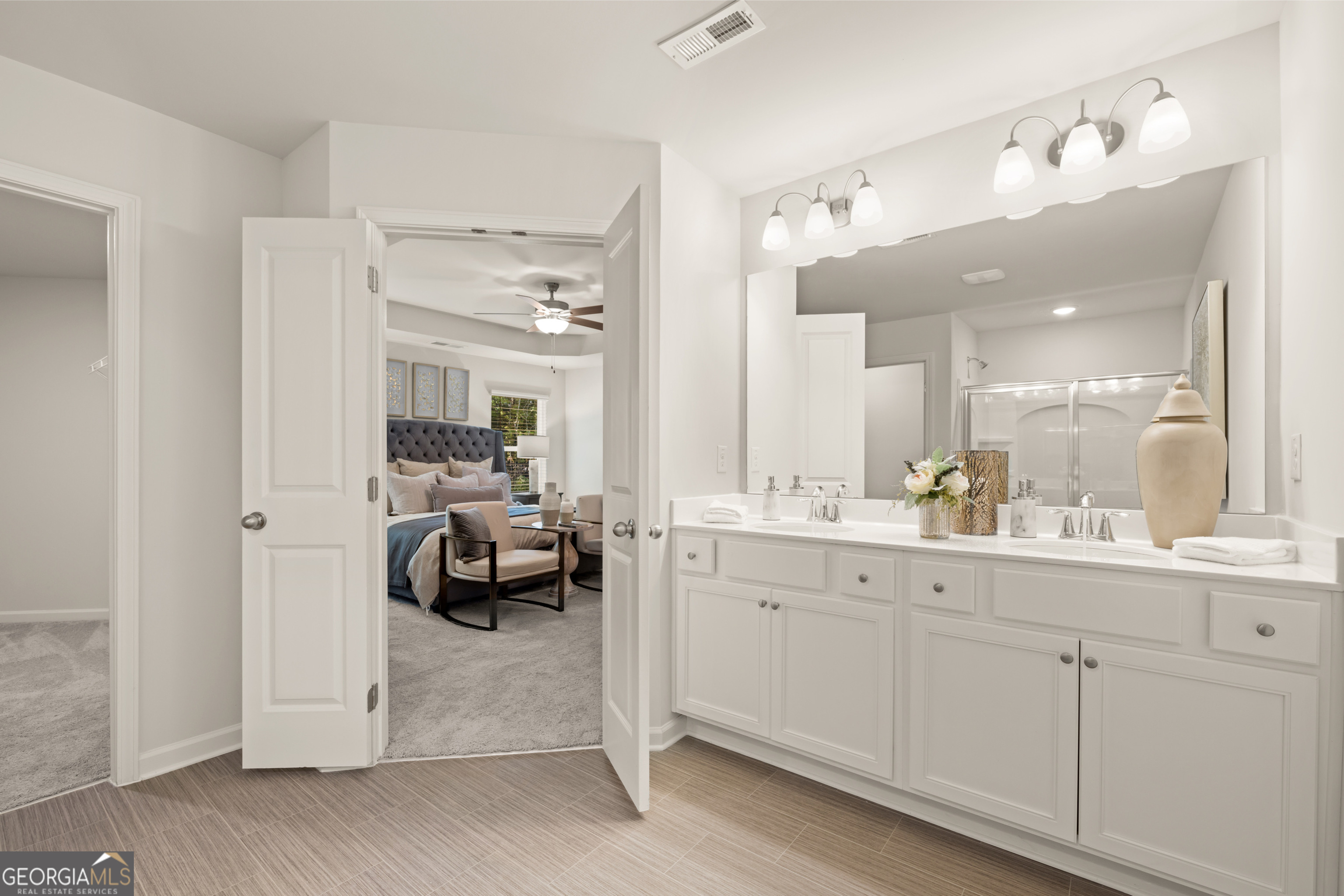 Staged bathroom with a long double-sink vanity and open doors to a carpeted bedroom.