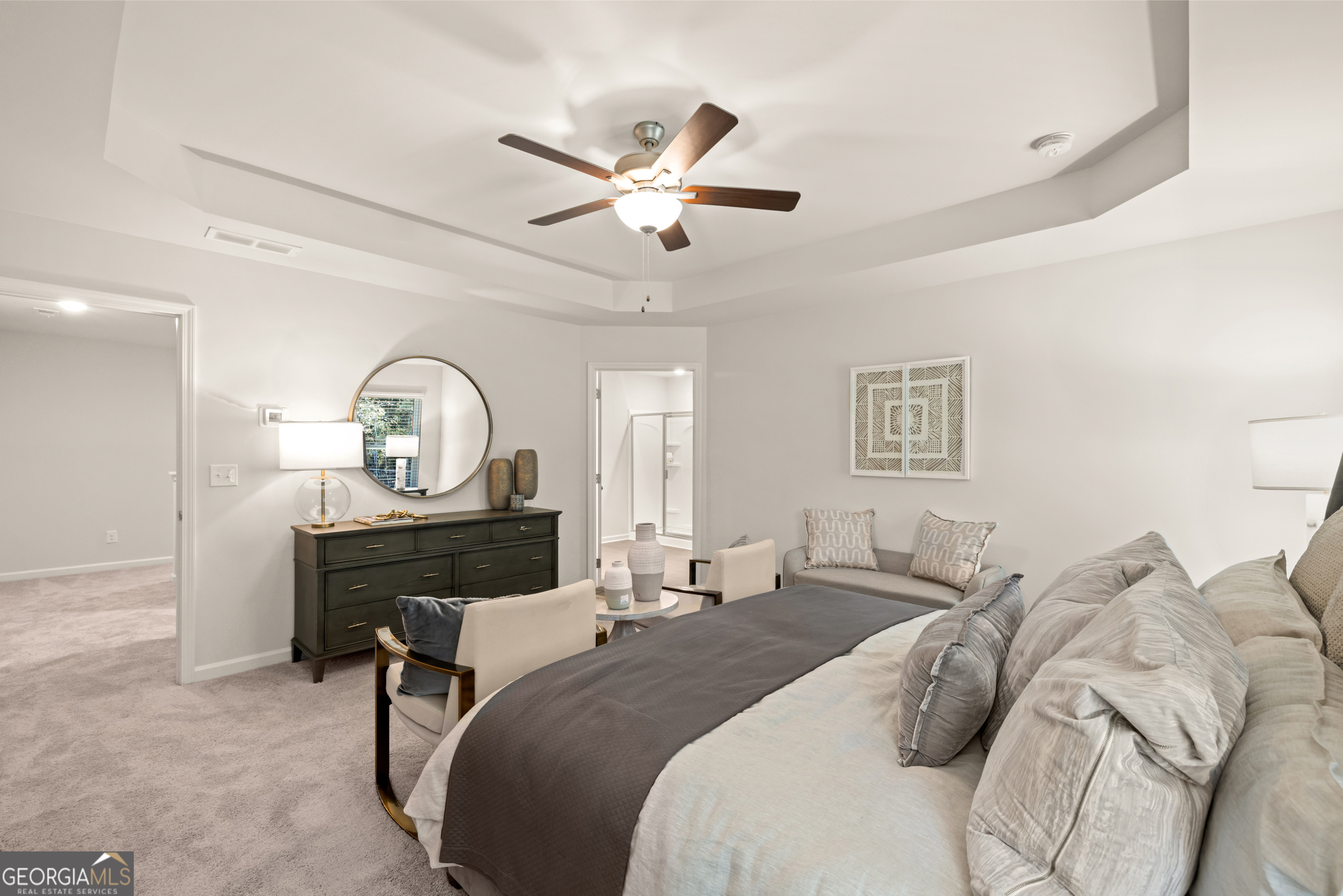 Staged primary bedroom with a tray ceiling, gray-and-white bed, dark dresser, and adjoining bathroom doorway.
