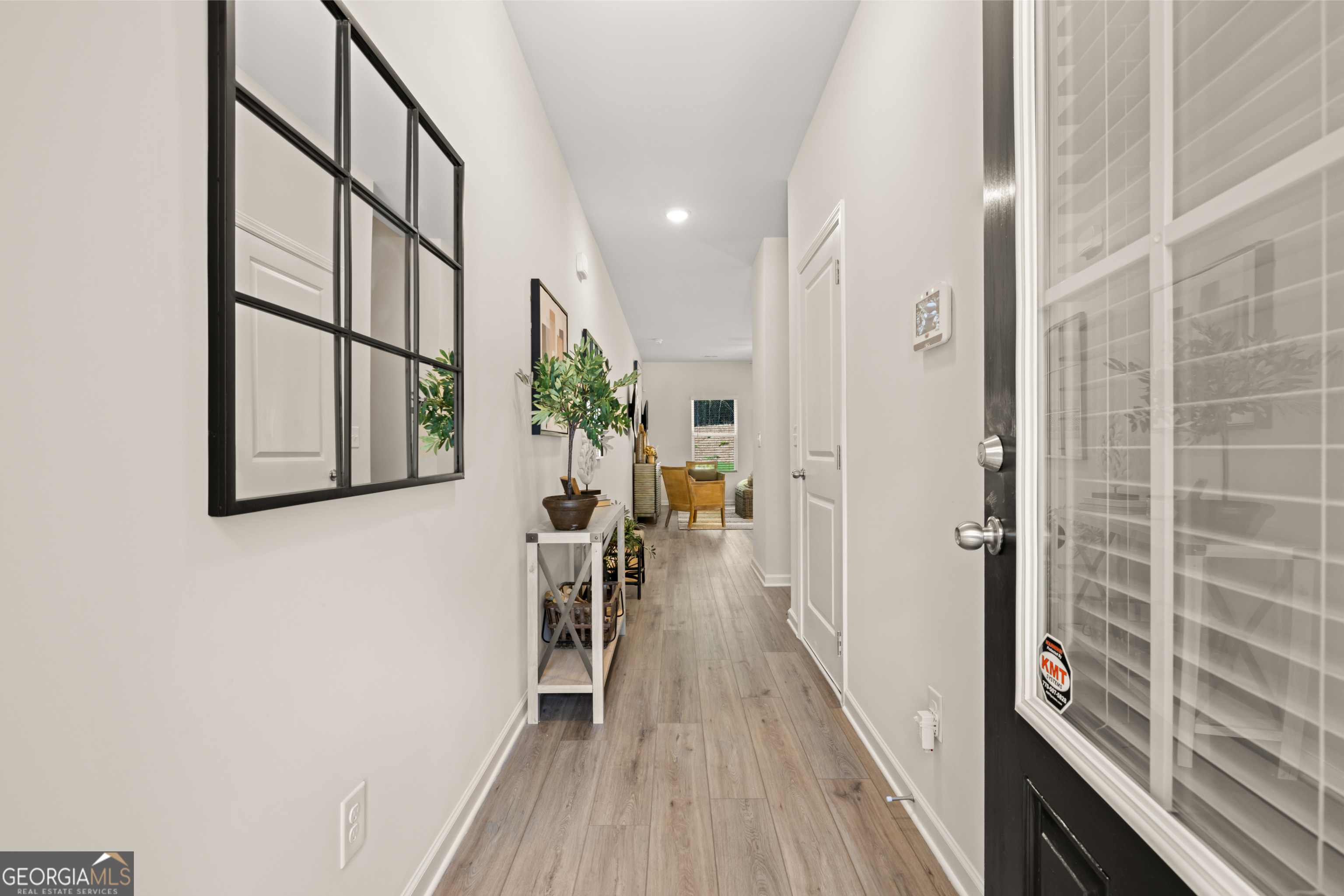 Staged home entry hallway with wood-look flooring, black-framed mirror, console table, and sitting area beyond.