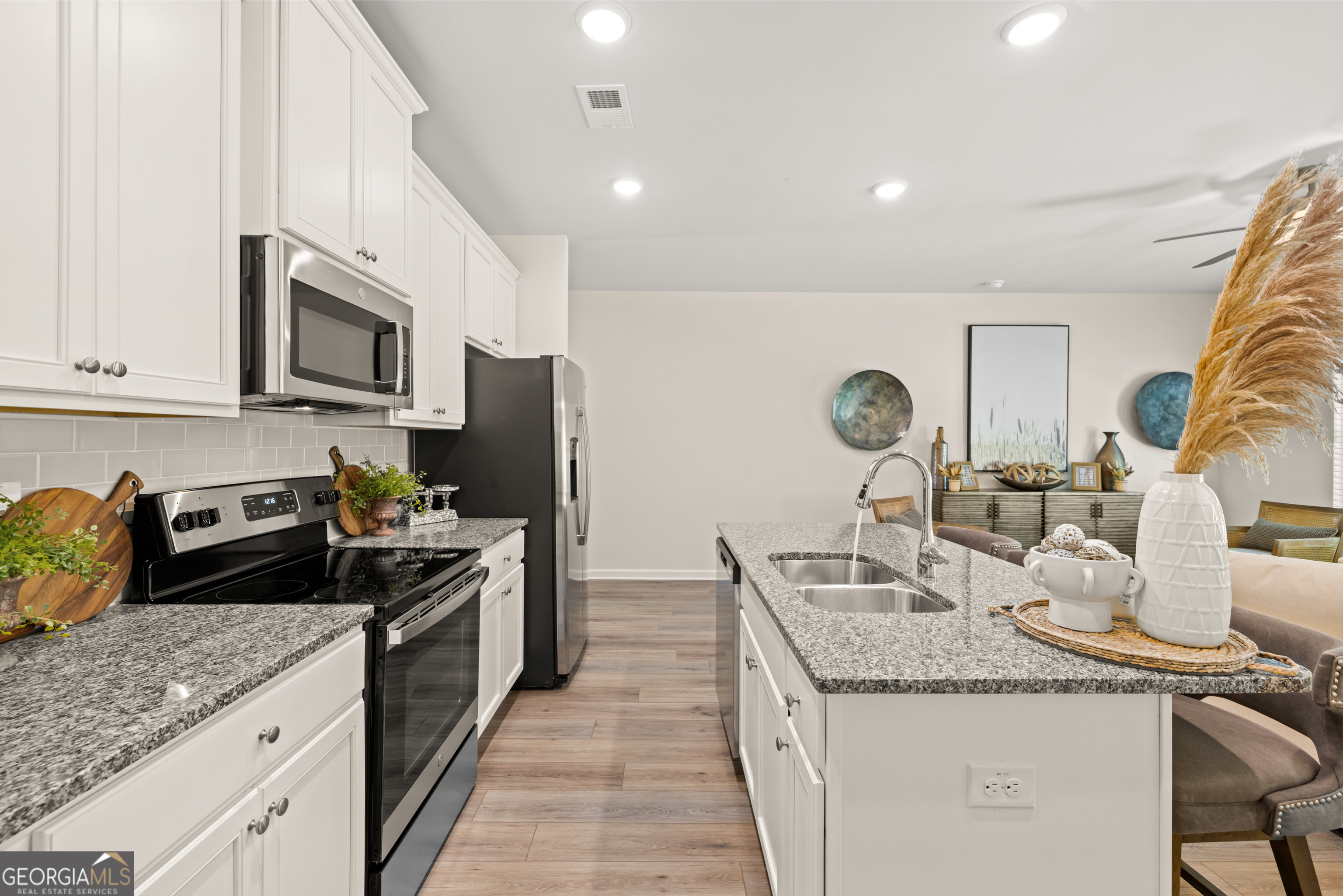 Staged kitchen with a granite island, double-basin sink, white cabinets, and stainless steel appliances.