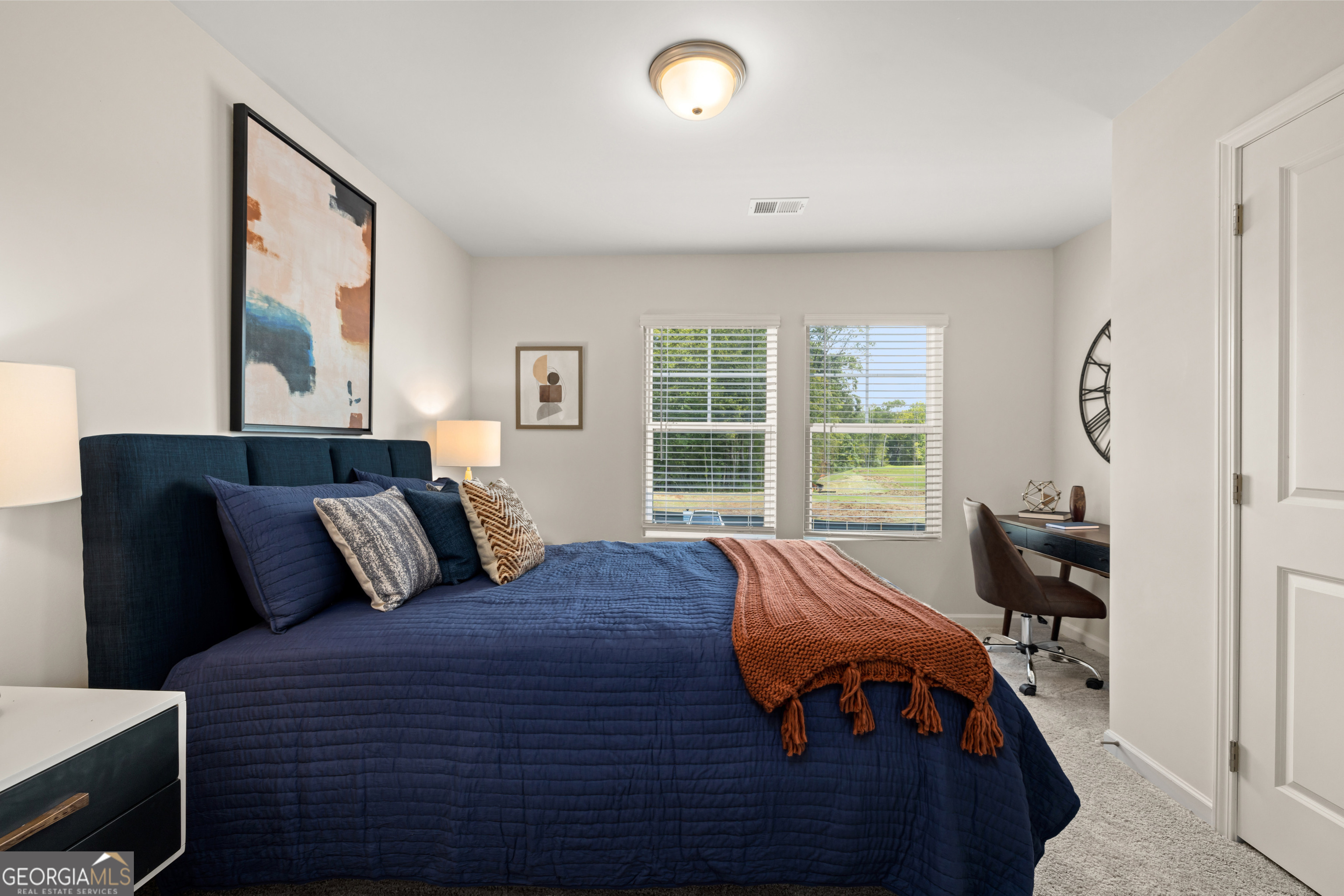 Staged bedroom with a navy bed, rust-colored throw, and two windows with white blinds.